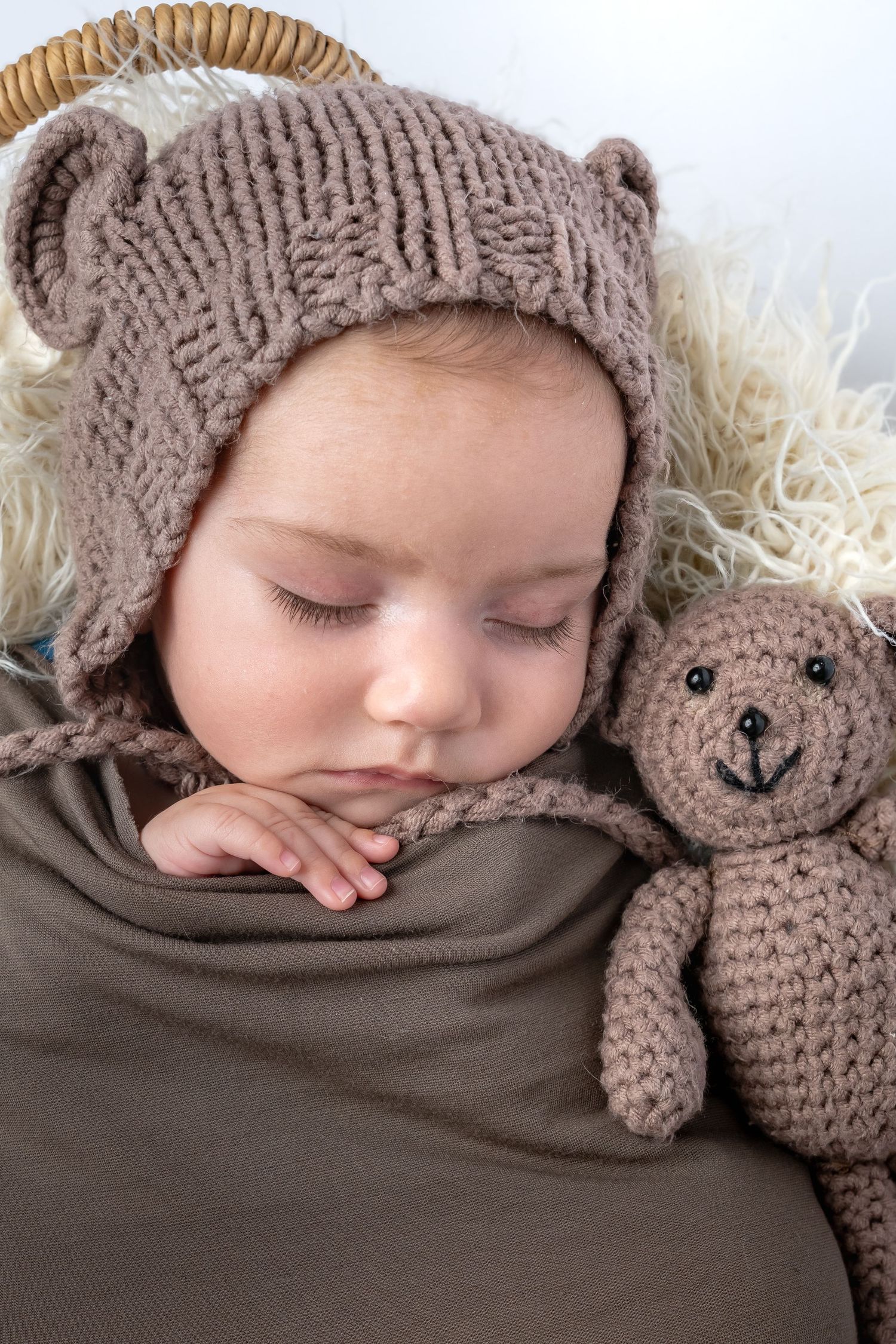 Sleeping newborn baby wearing a knit bear ear bonnet hat with a matching crocheted teddy bear toy.