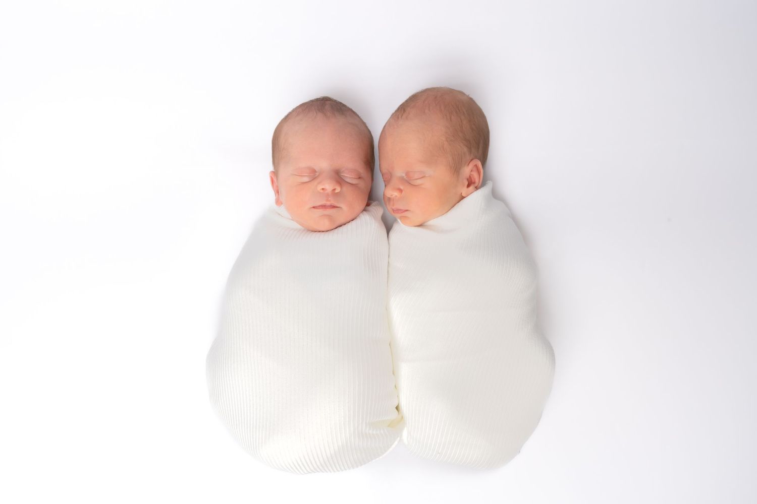 Two swaddled newborn twins sleeping peacefully side by side in white wraps on a white background.