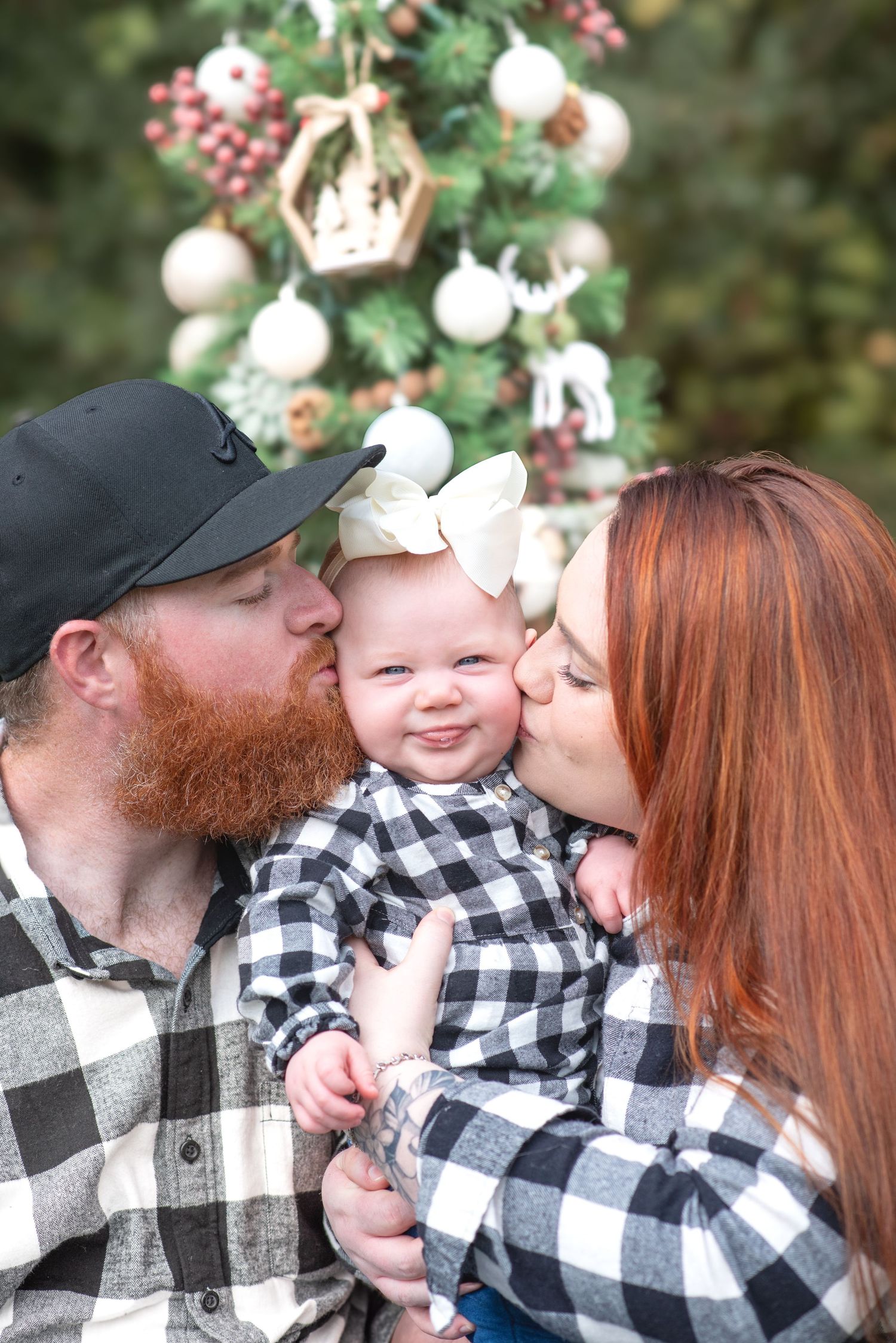 A happy family with a bearded dad and redhead mom kissing their smiling baby girl in front of a Christmas tree.