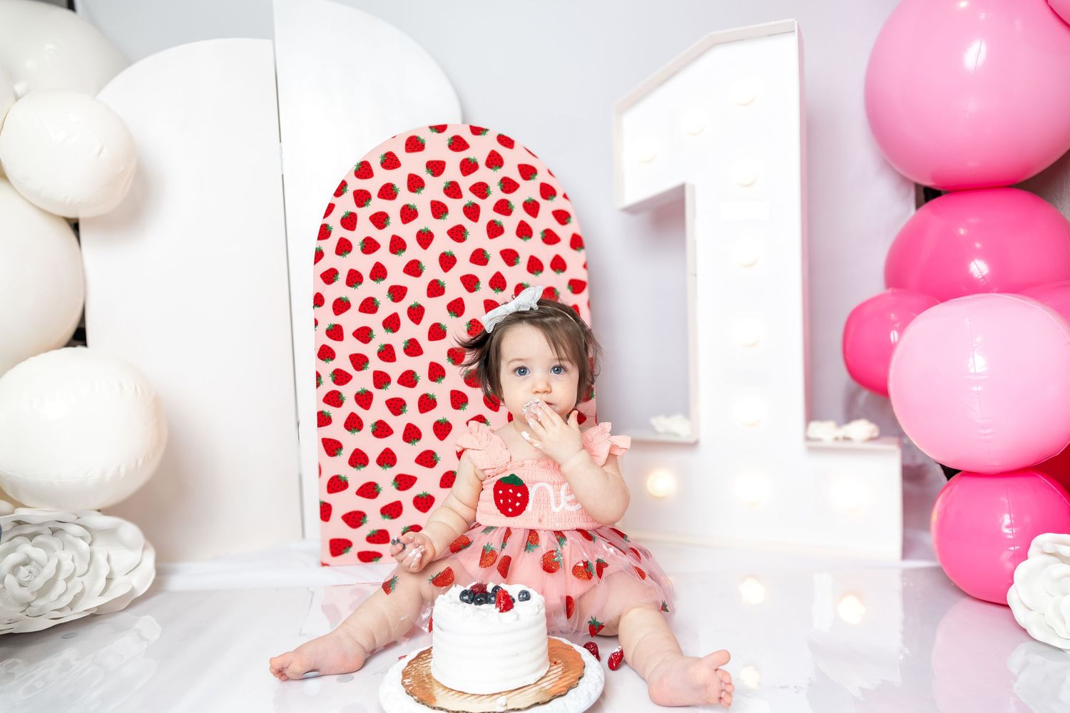 Baby girl in strawberry dress celebrates first birthday with cake, surrounded by pink balloons and number one decorations.
