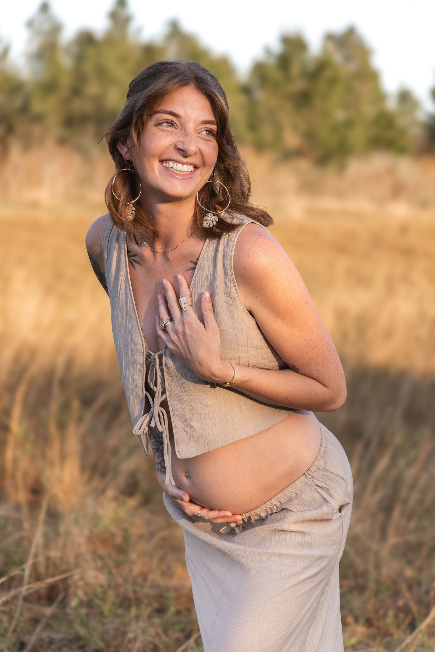 Smiling pregnant woman in beige dress holding baby bump outdoors in golden field at sunset.