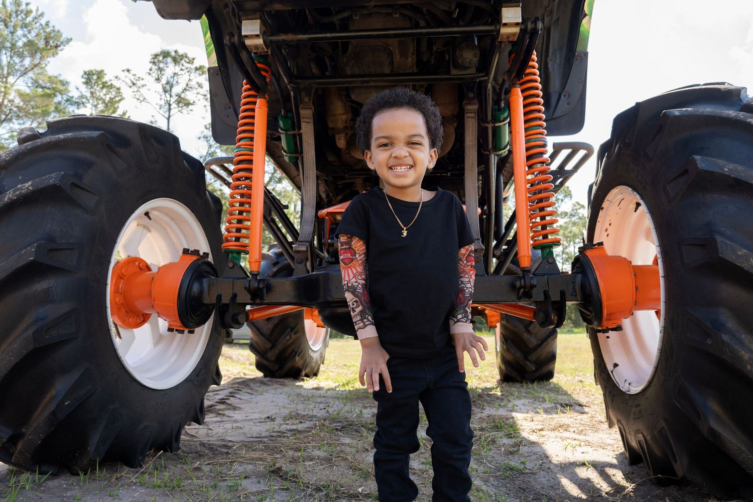 Smiling child standing between giant monster truck tires with orange accents outdoors.