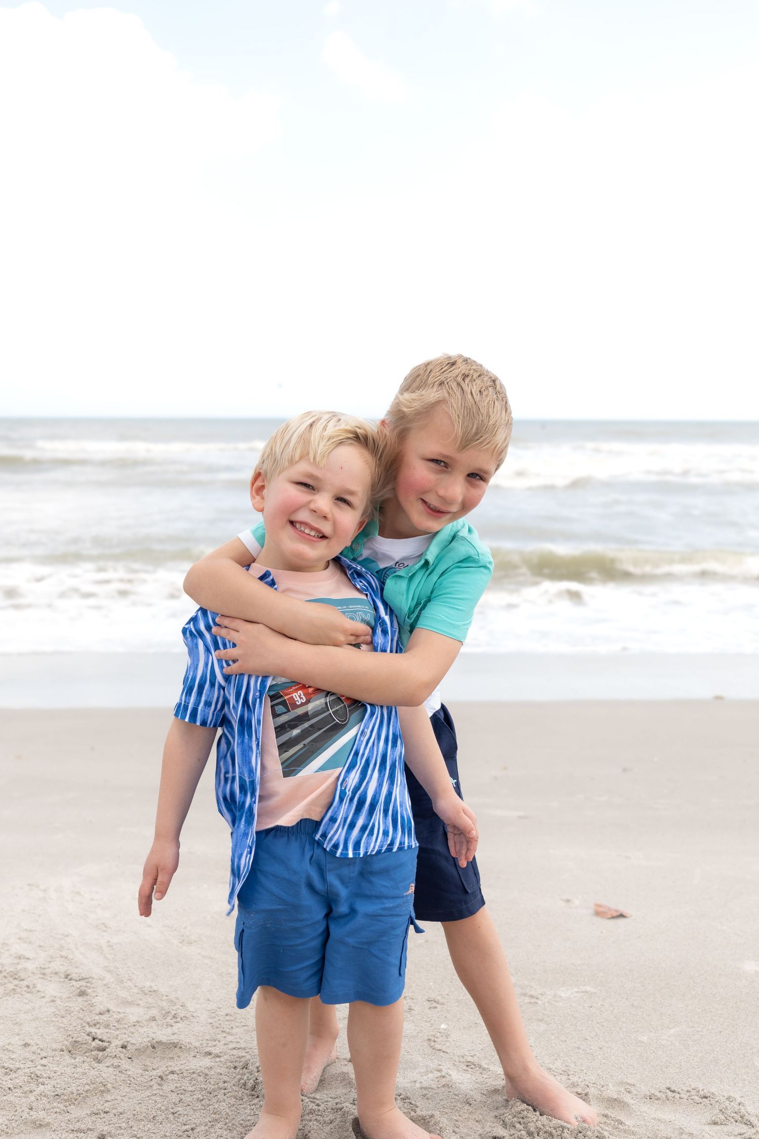 Two young brothers share a hug on a sandy beach with ocean waves in the background.