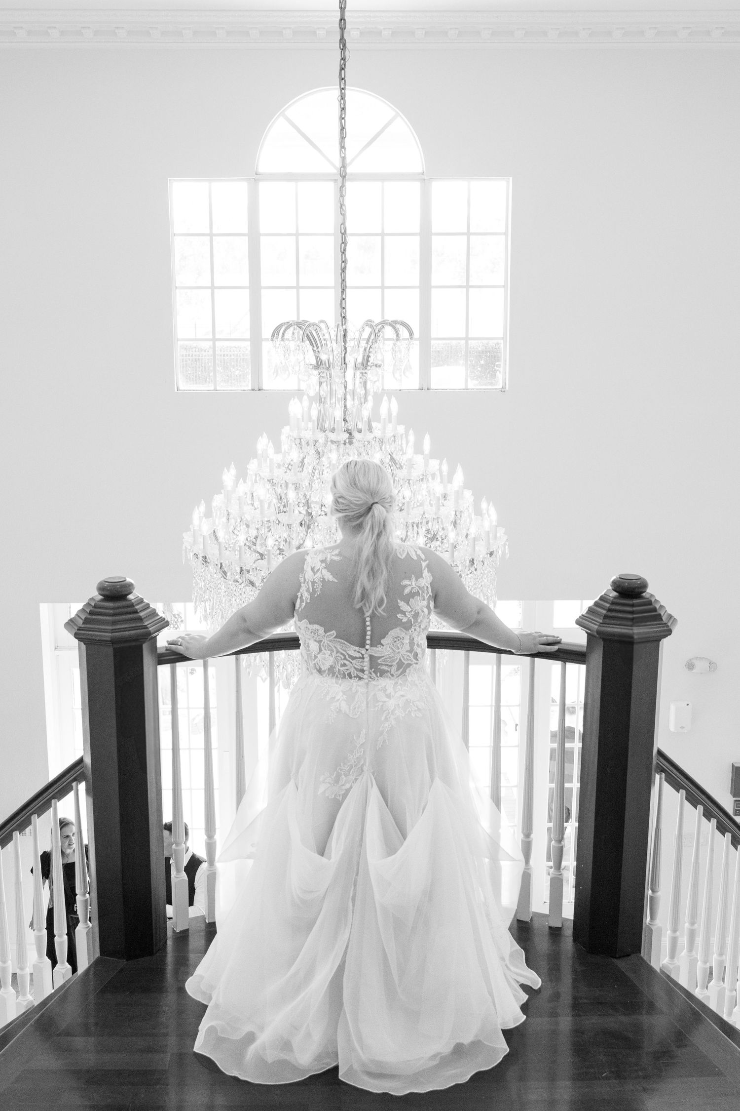 Bride in lace wedding gown stands at elegant staircase railing beneath crystal chandelier in black and white photo.