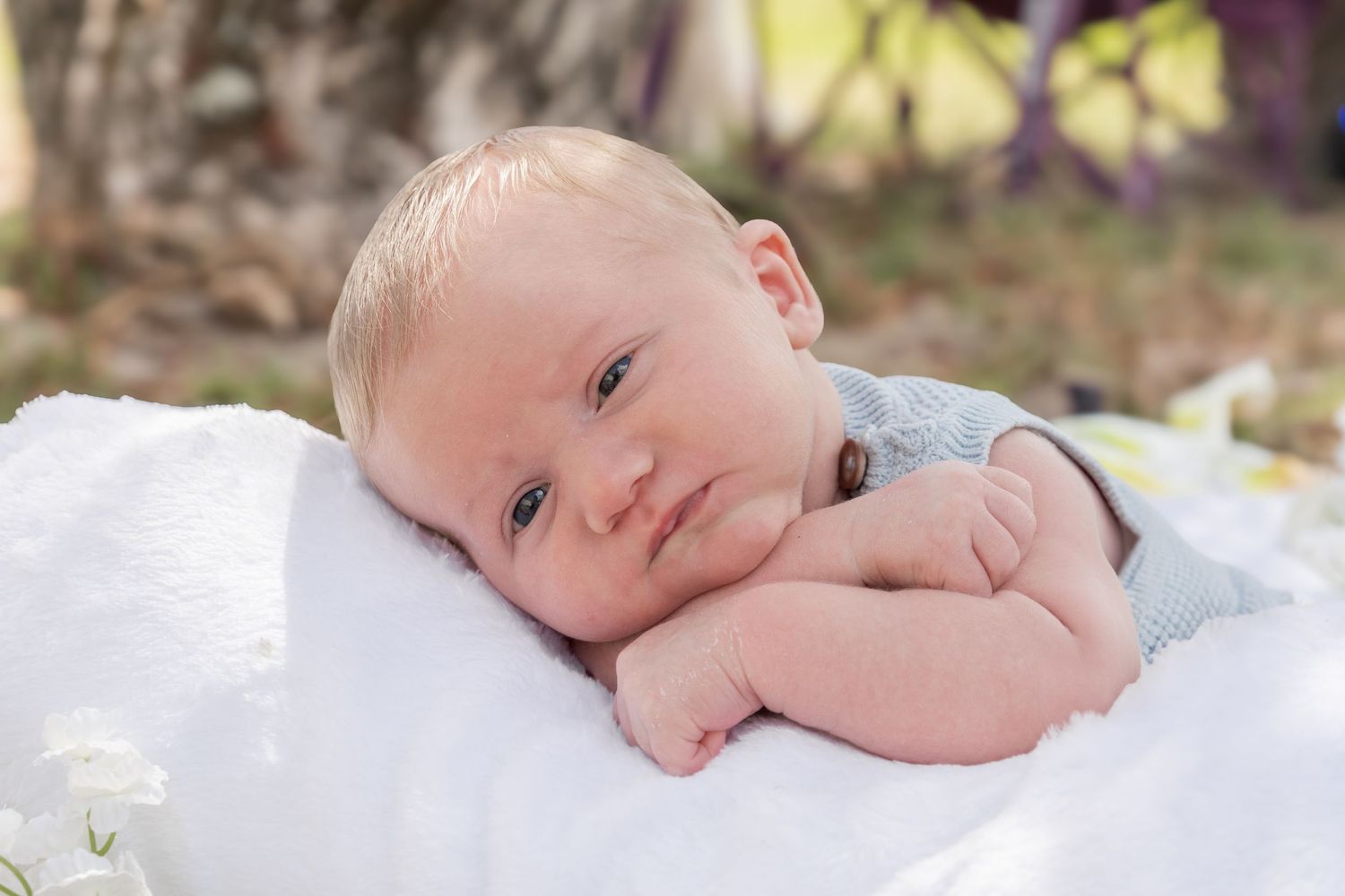 Newborn baby lying on white blanket outdoors, resting chin on hands with soft natural background.