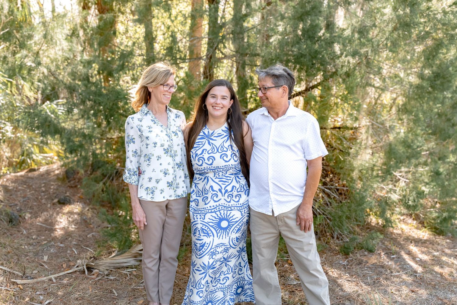 Family of three smiling together outdoors among tall pine trees in a sunny forest setting.