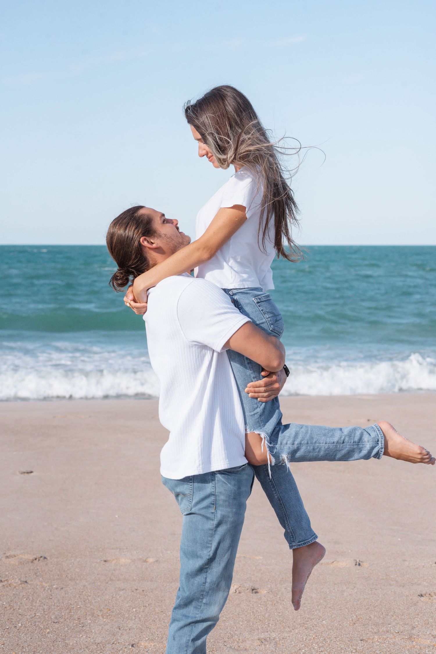 Couple in matching white shirts and jeans embrace on a sunny beach with ocean waves in the background.