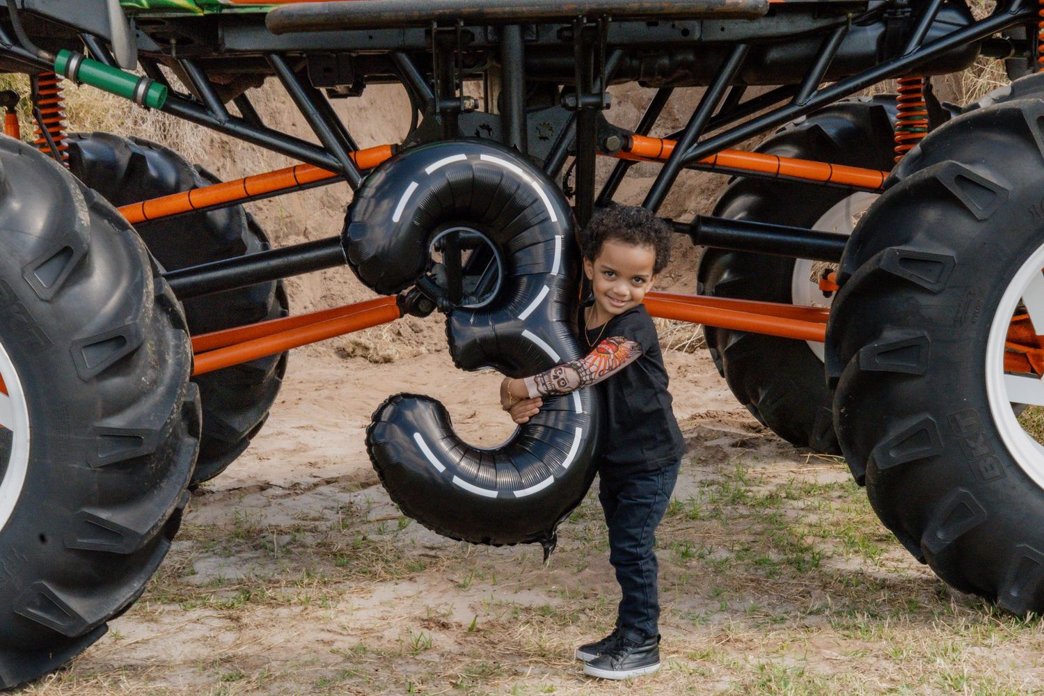 A child holds a black number 3 balloon beside a massive monster truck at an outdoor birthday celebration.