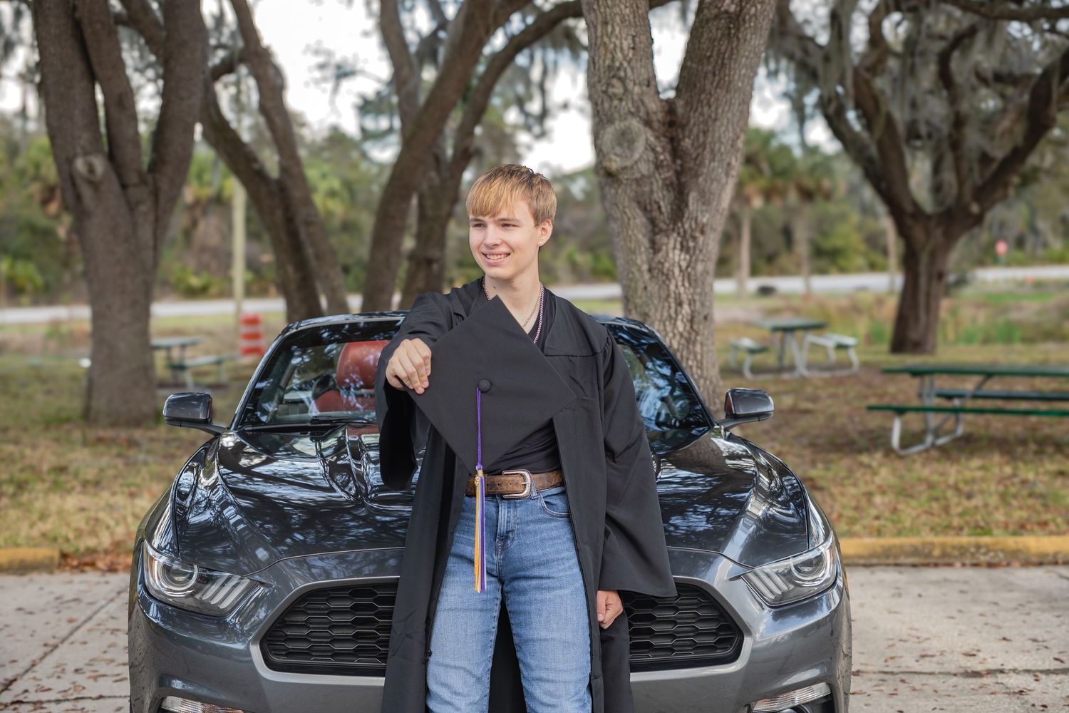 Short-haired woman in black blazer and jeans posing beside a gray Ford Mustang in a tree-lined parking area.