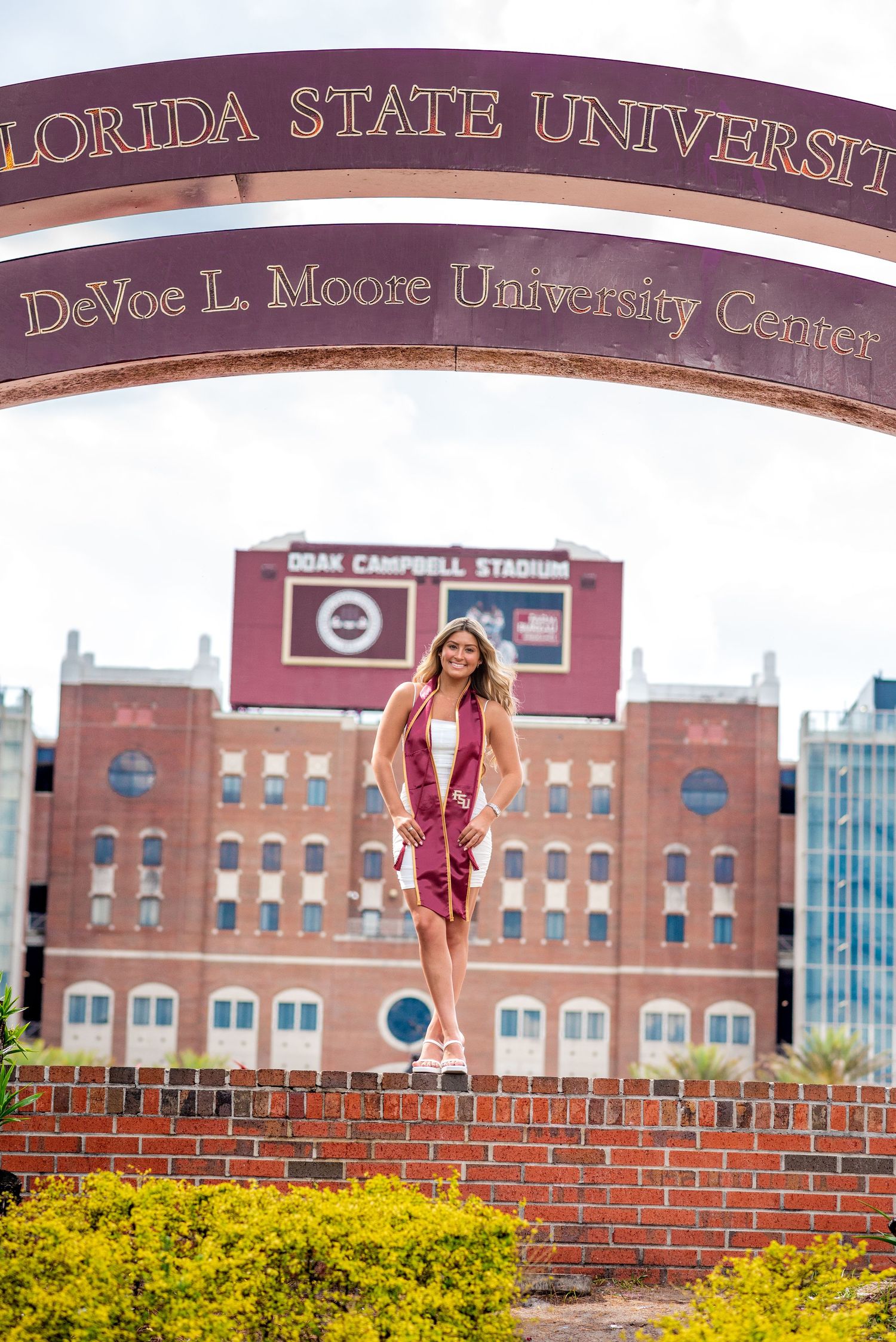 FSU graduate in cap and gown poses under Florida State University arch at DeVoe L. Moore University Center.