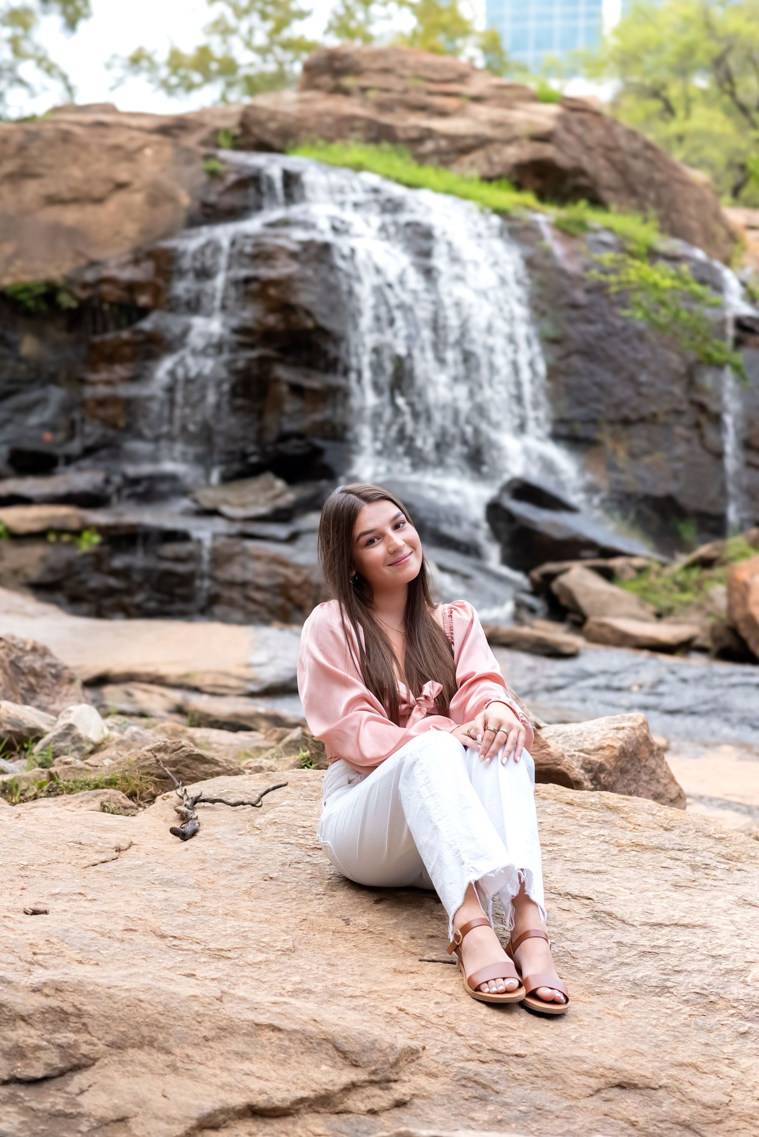Young woman in pink blouse and white jeans sitting on rocks in front of a cascading waterfall outdoors.