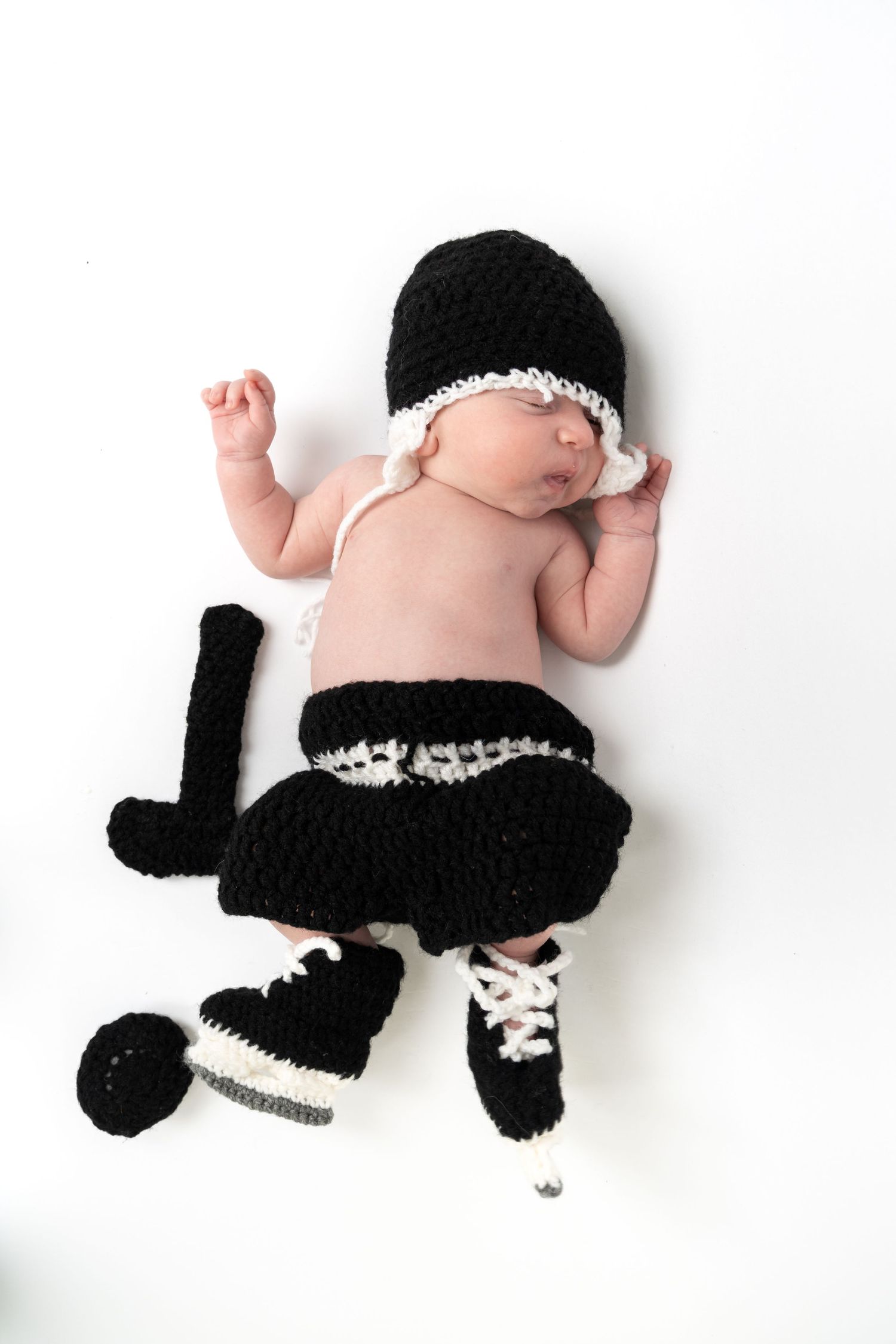 Sleeping newborn baby in black and white crocheted hockey player costume with skates and stick on white background.