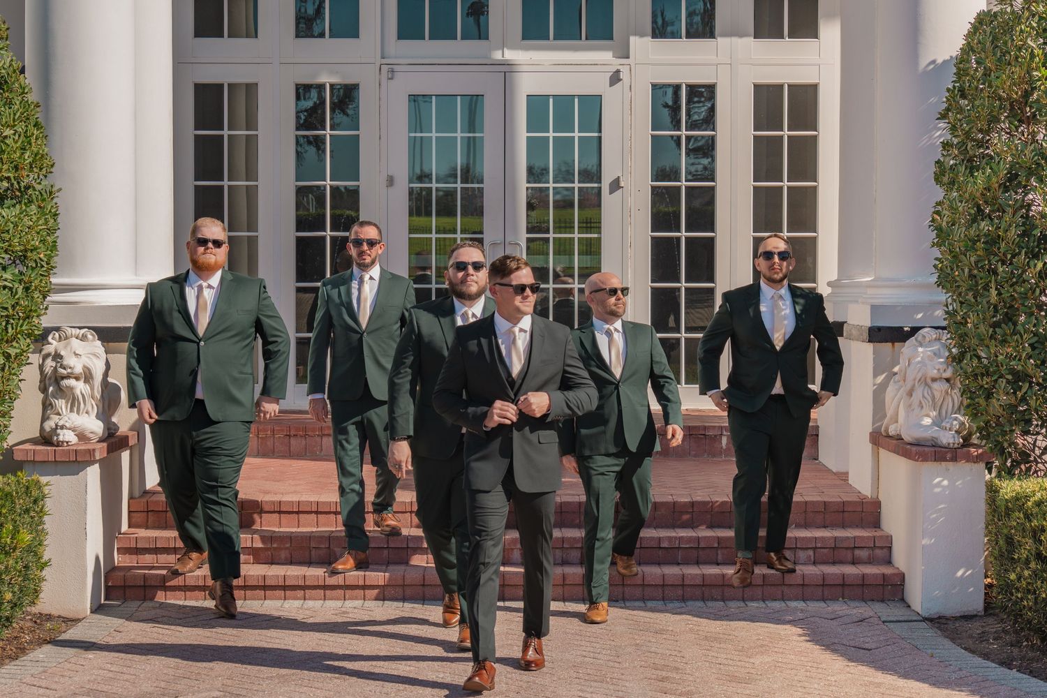 Groomsmen in dark green suits and sunglasses walk confidently down brick steps outside an elegant white venue.