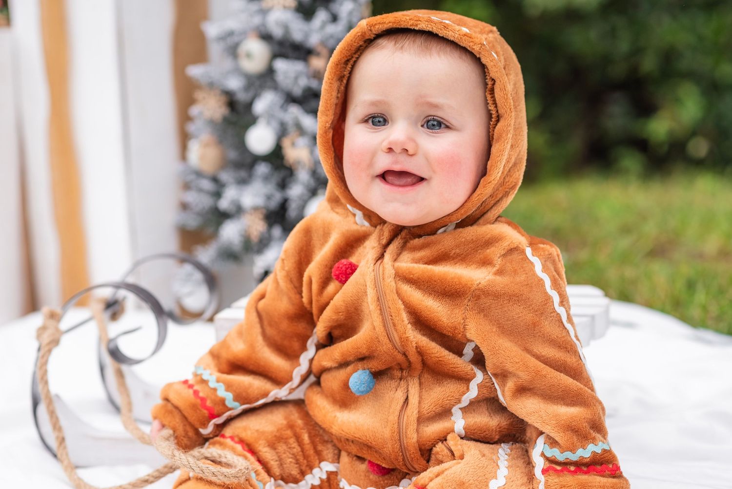 Adorable baby in a gingerbread man costume sitting in front of a Christmas tree outdoors.