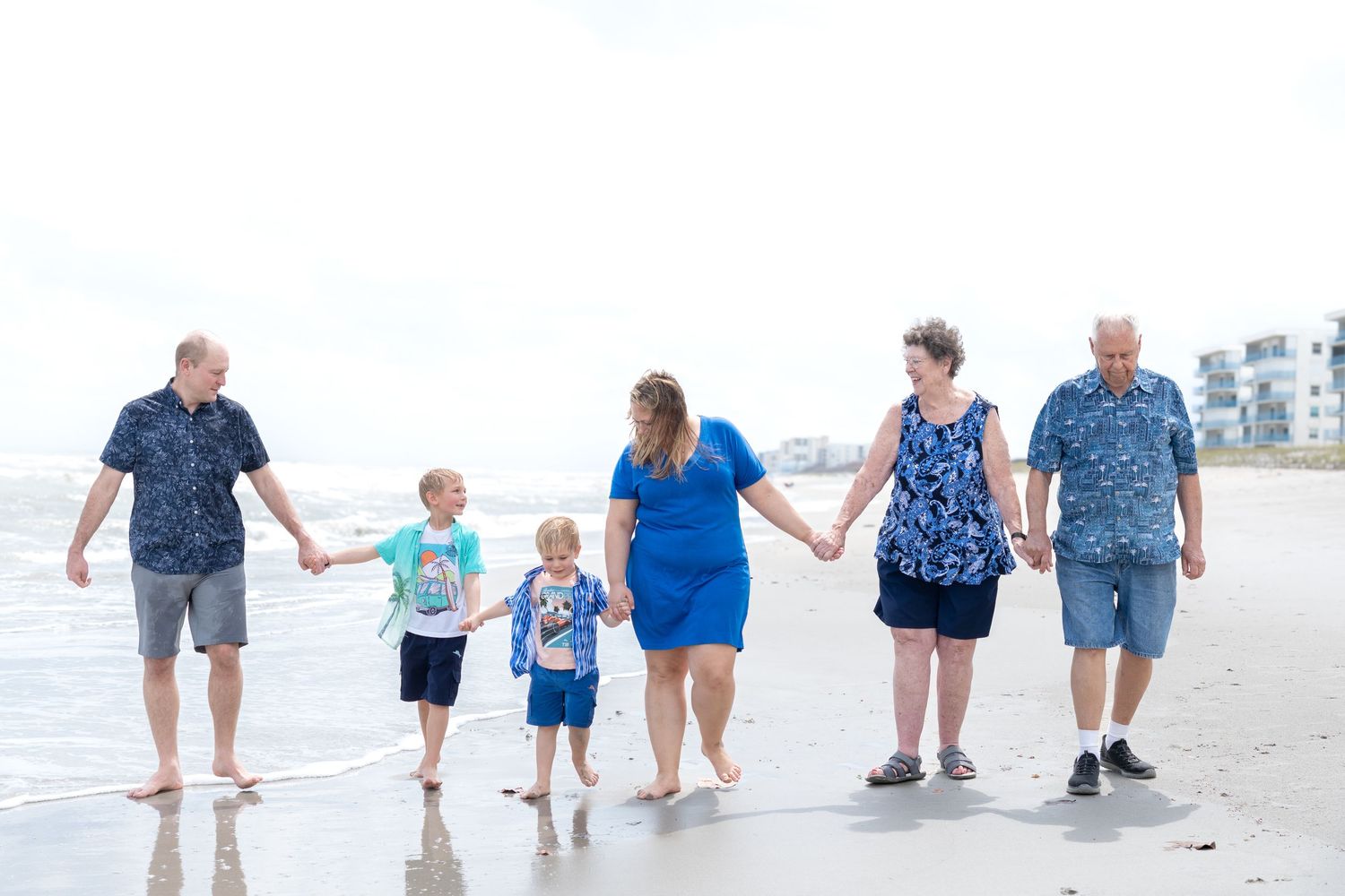 Multi-generational family holding hands walking along a sandy beach on a cloudy day near the ocean.
