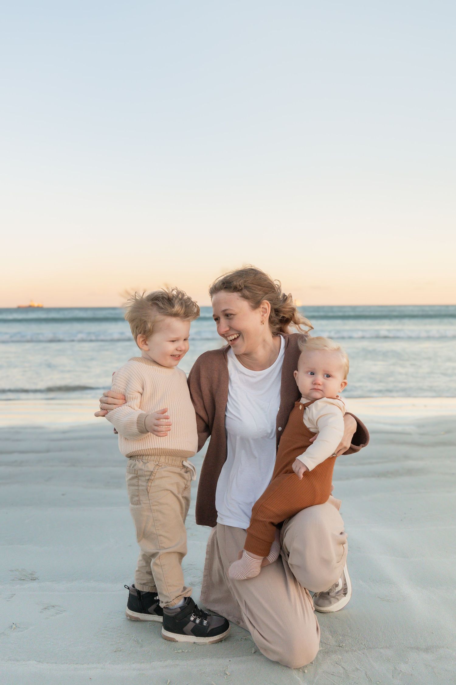 A smiling mother kneels on a sandy beach at sunset, holding a baby while embracing a toddler beside her.