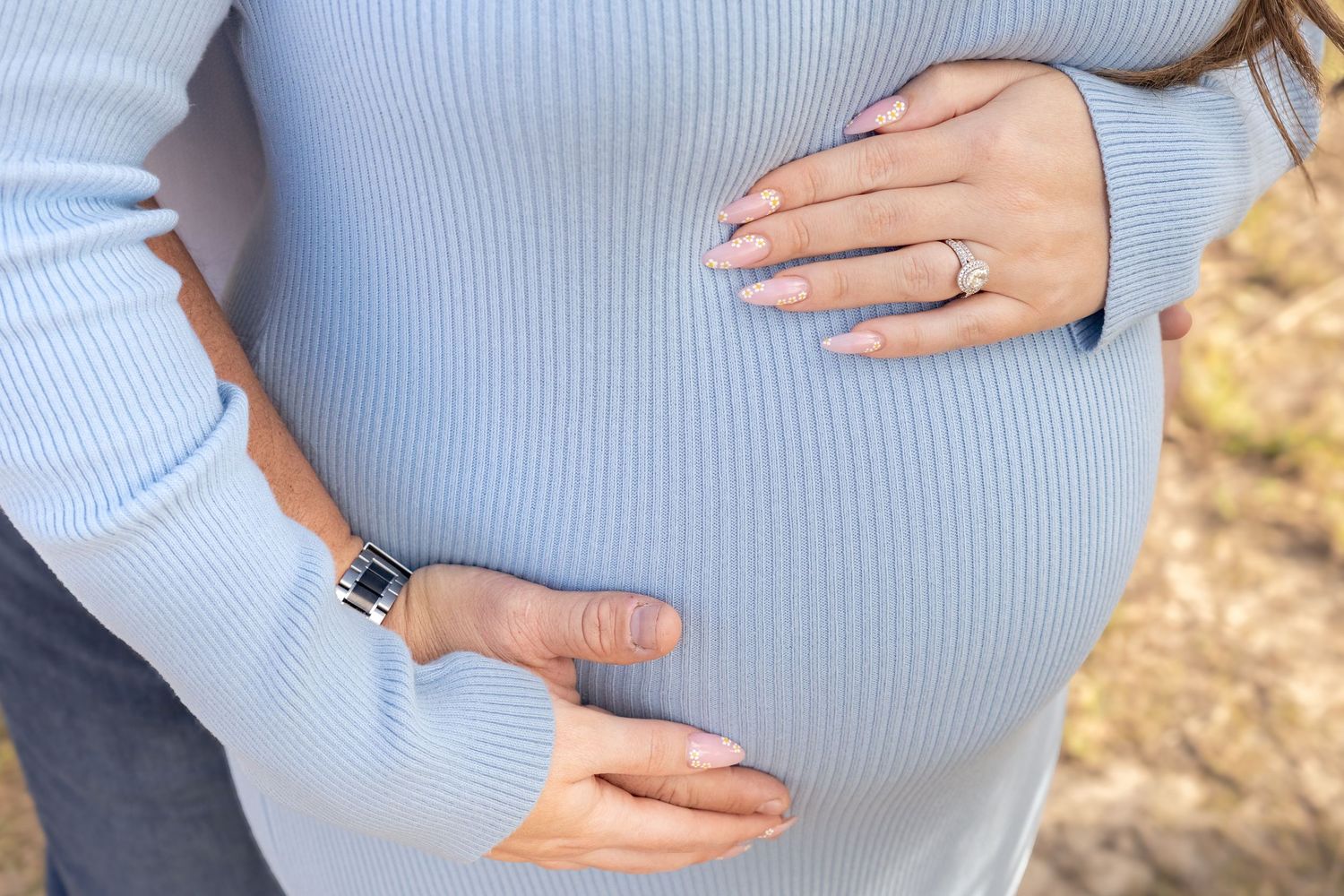 Close-up of pregnant woman in blue sweater with partner's hands cradling baby bump outdoors.
