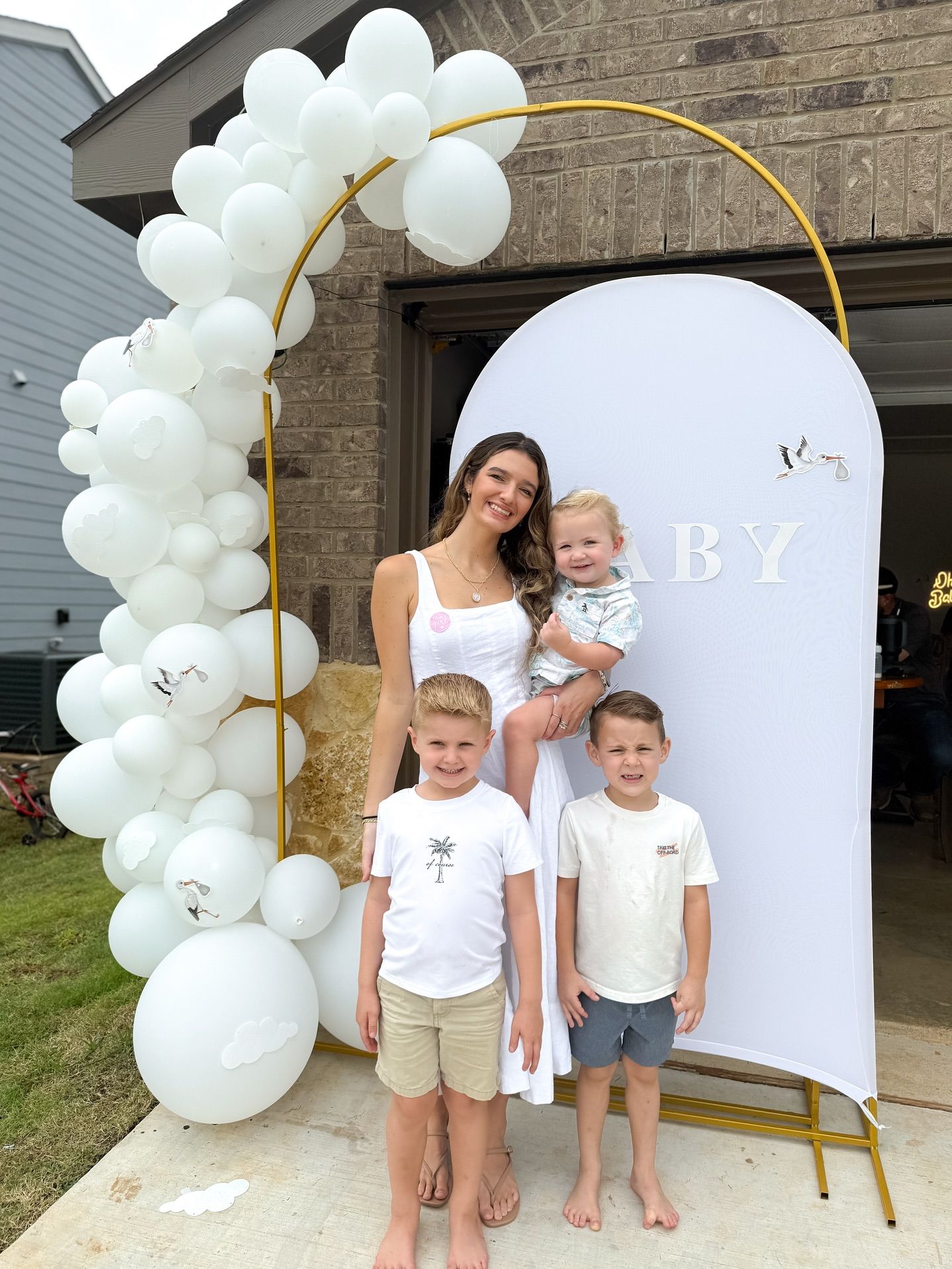 Woman in white dress holding baby with two young boys posing at baby shower balloon arch display.