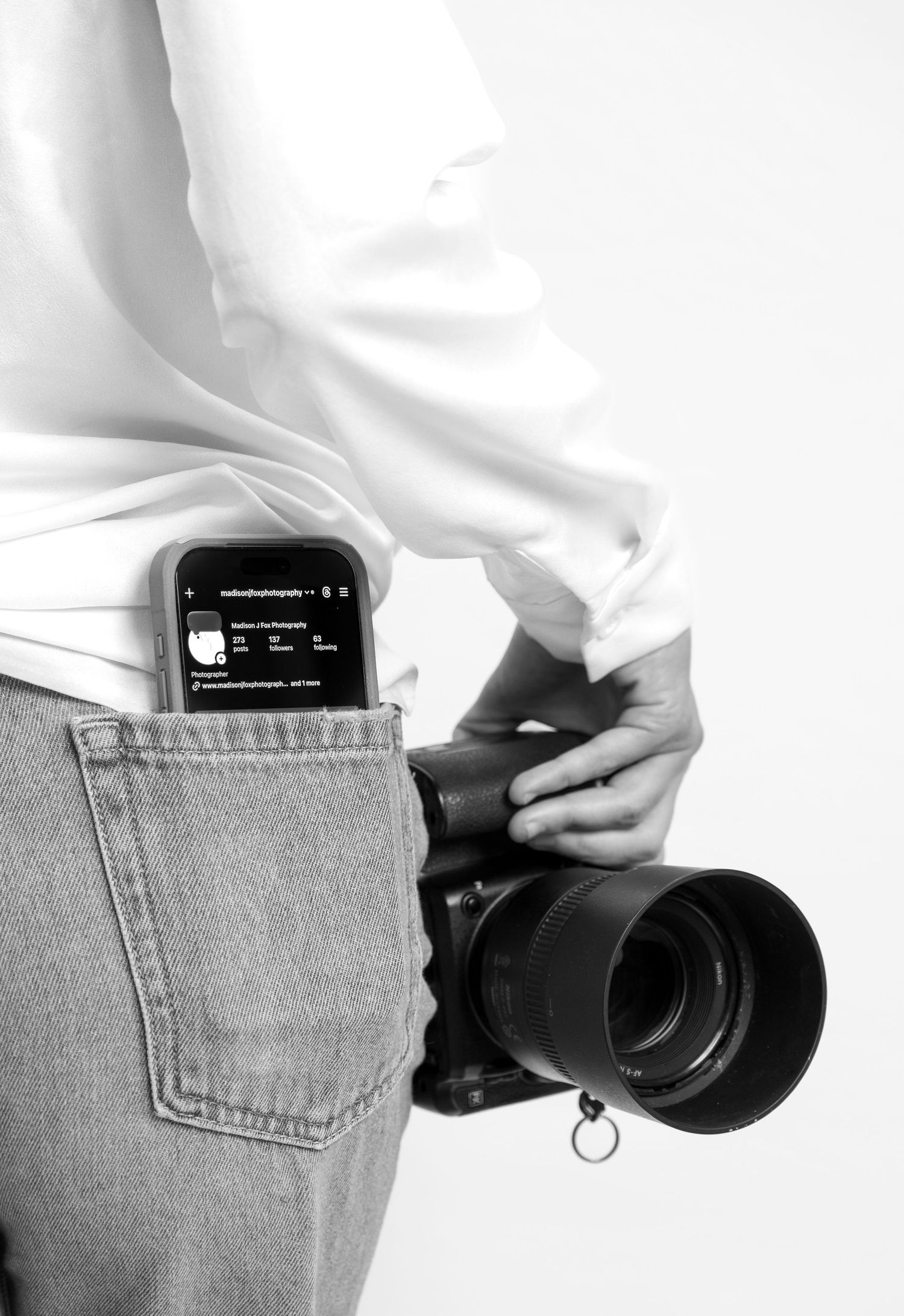 Black and white close-up of photographer holding DSLR camera with phone showing Instagram profile in pocket.