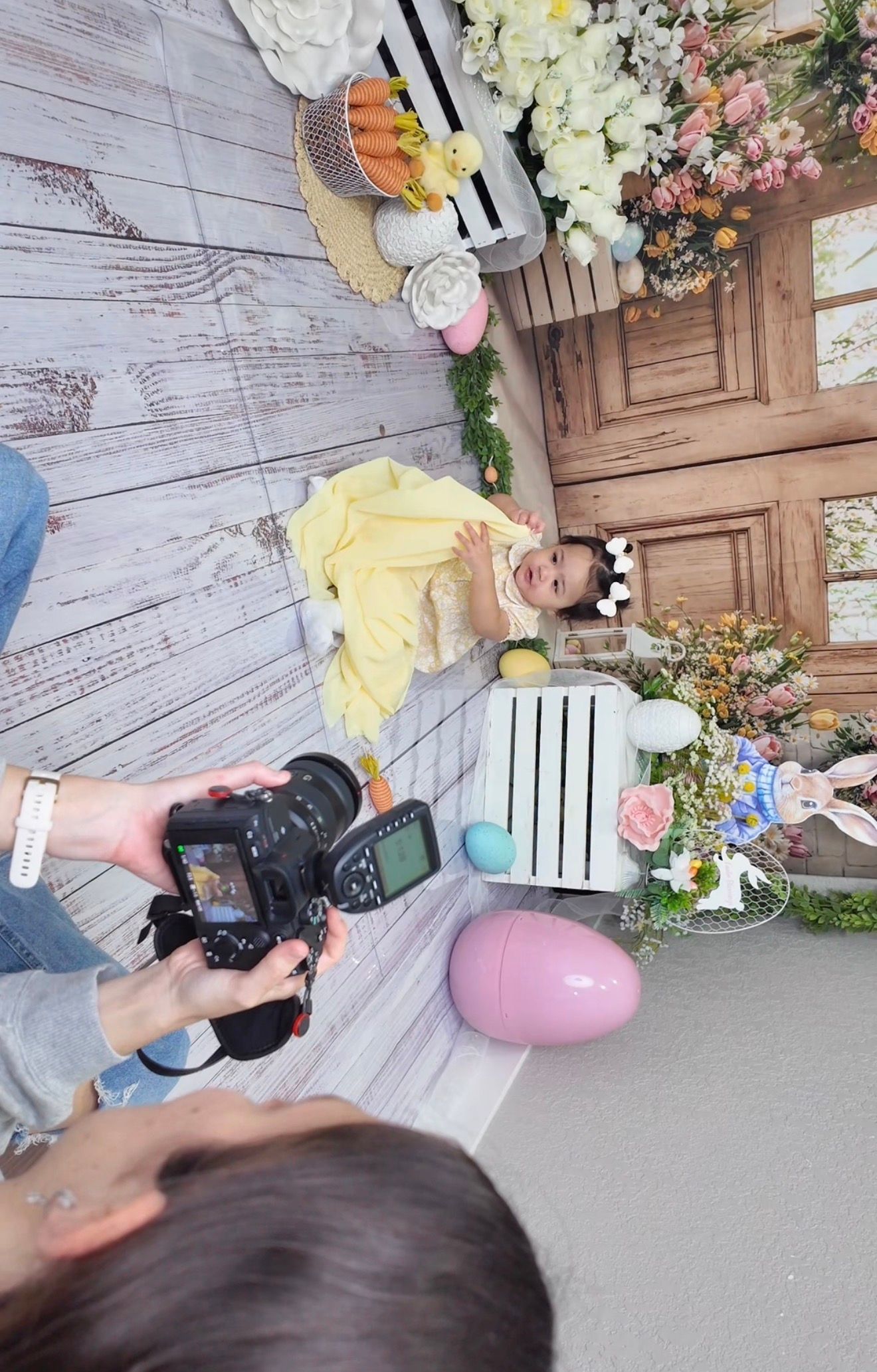 Photographer captures overhead shot of baby in yellow dress during Easter-themed photo session with floral decorations.