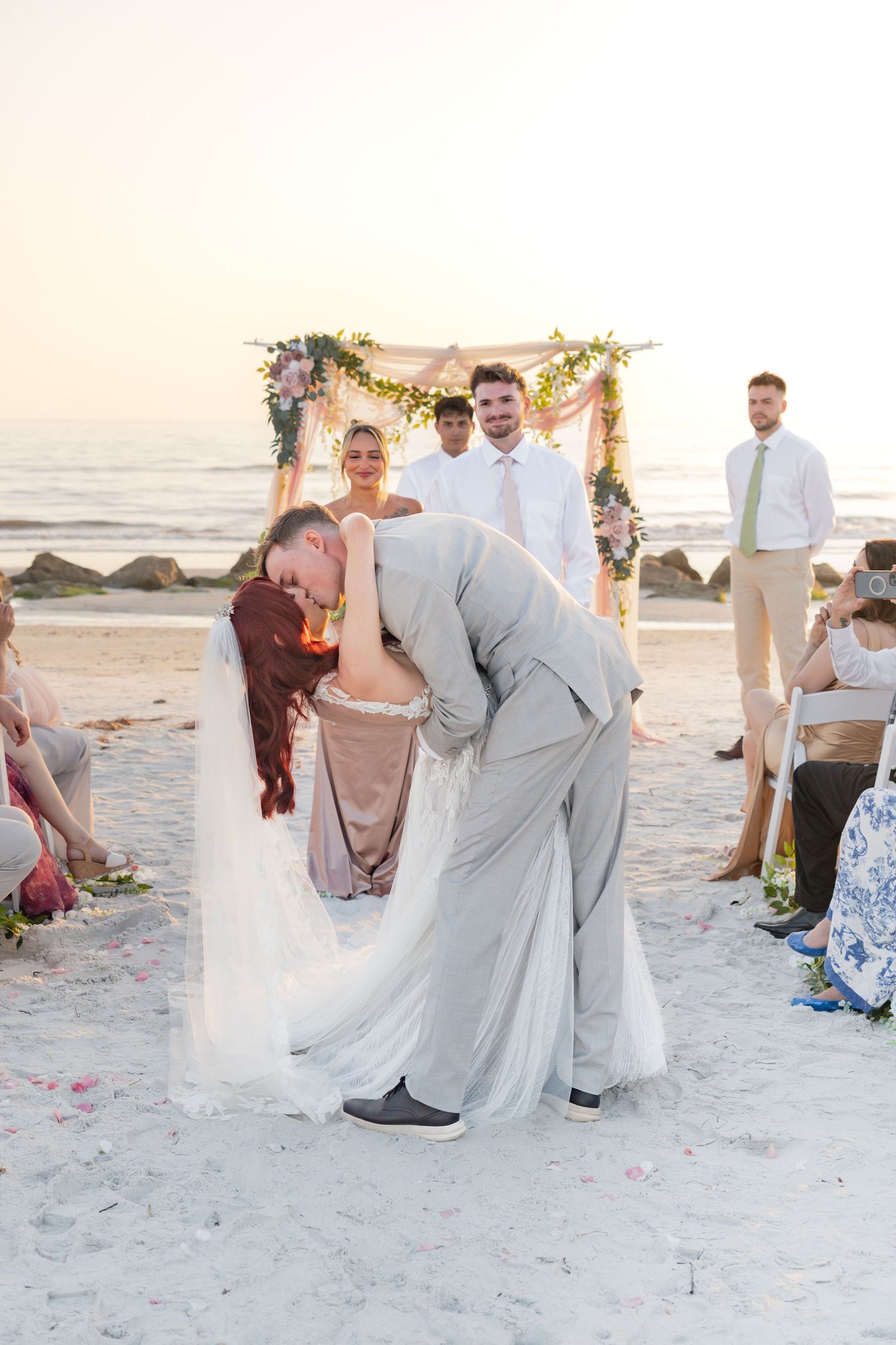 Groom dips bride for a romantic kiss on a sandy beach at sunset during outdoor wedding ceremony.