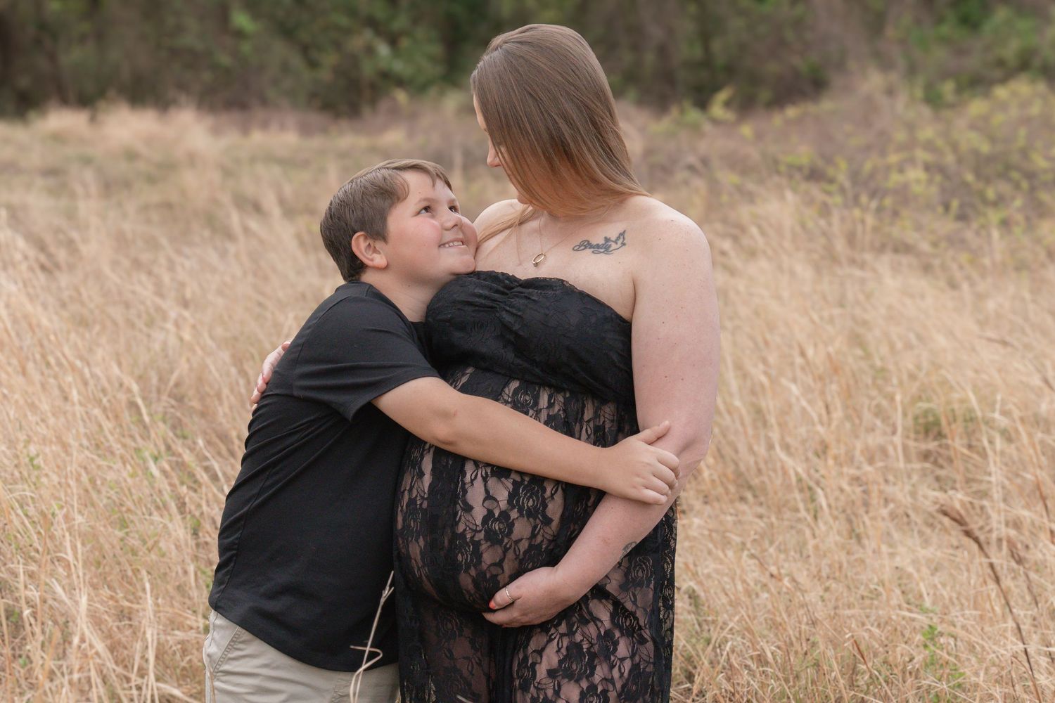Couple embracing in golden field, pregnant woman in black lace dress with tattoos, partner kissing baby bump outdoors.
