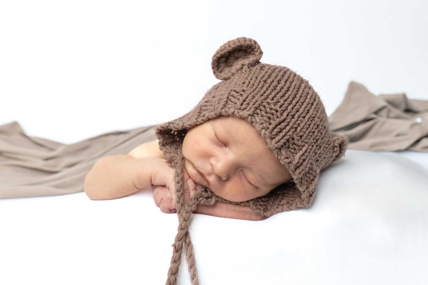 Sleeping newborn baby wearing a cozy brown knitted bear ear bonnet hat, posed on a soft white backdrop.