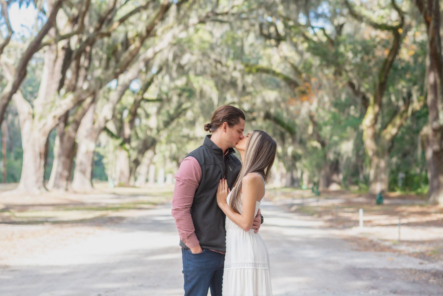 Couple sharing a romantic kiss on a tree-lined path draped with Spanish moss in a serene outdoor setting.