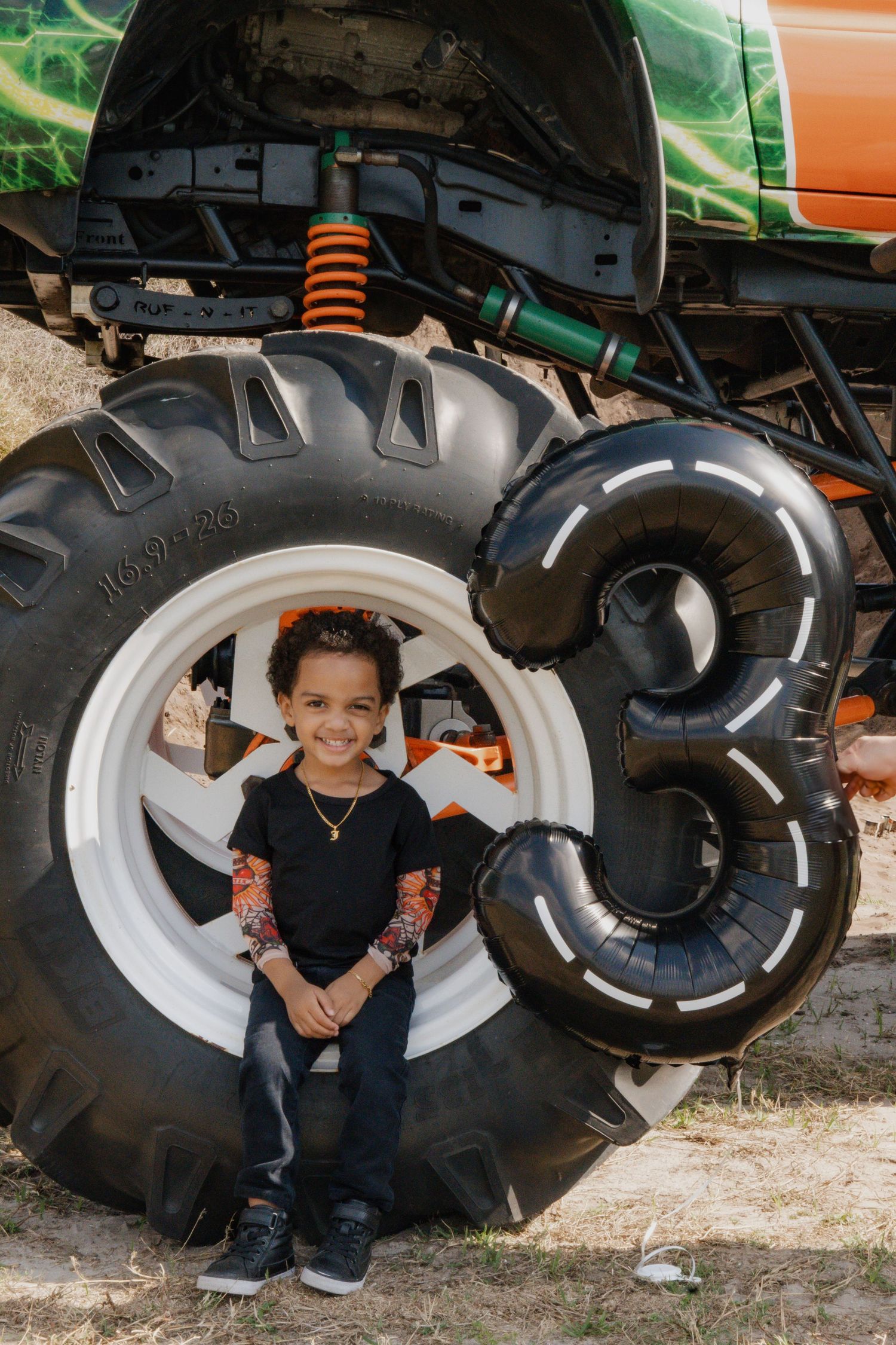 A young boy sits in front of a giant monster truck tire next to a number 3 balloon at his 3rd birthday party.