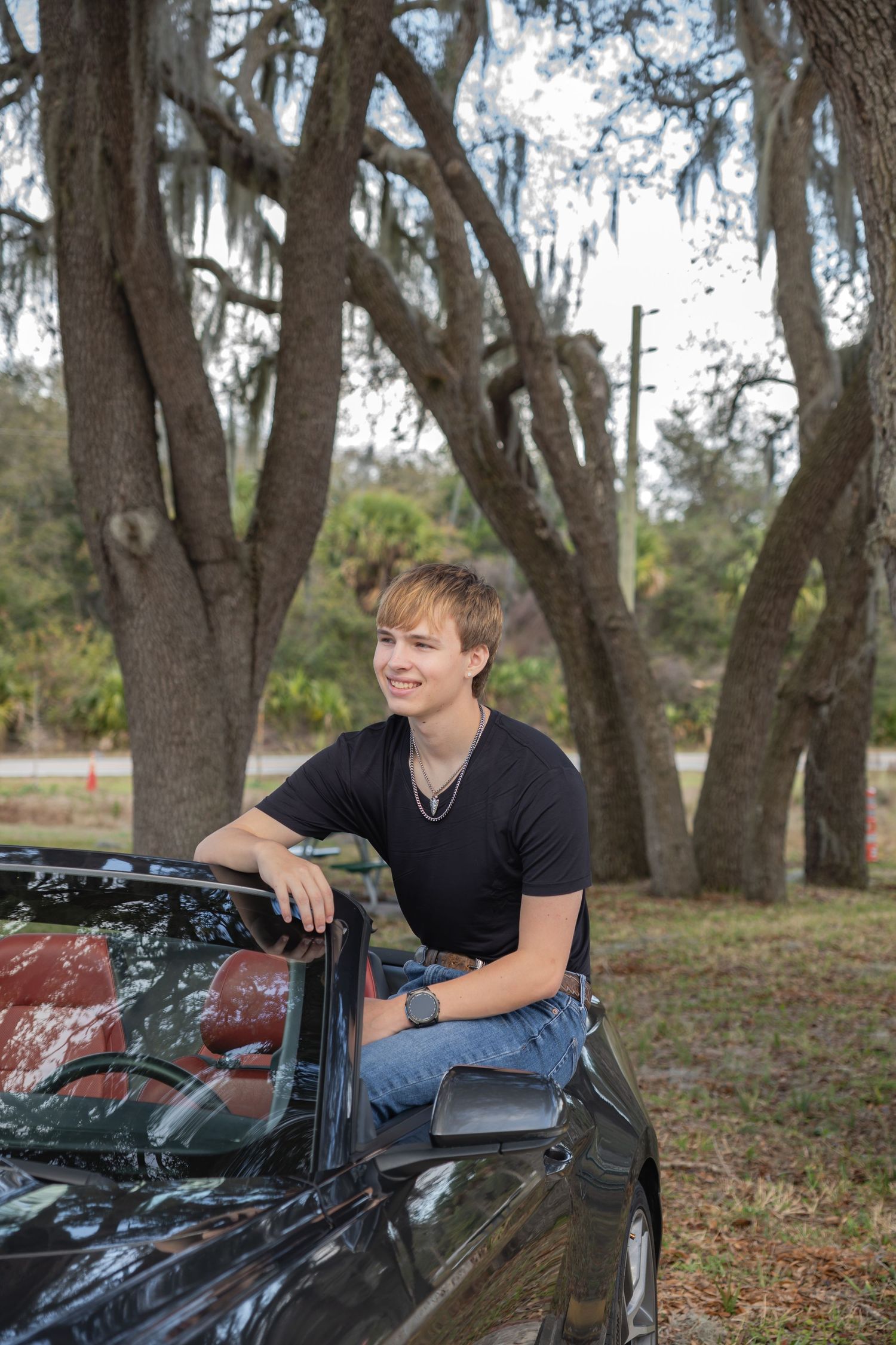 Smiling teen boy leaning on a black convertible car surrounded by moss-draped trees.