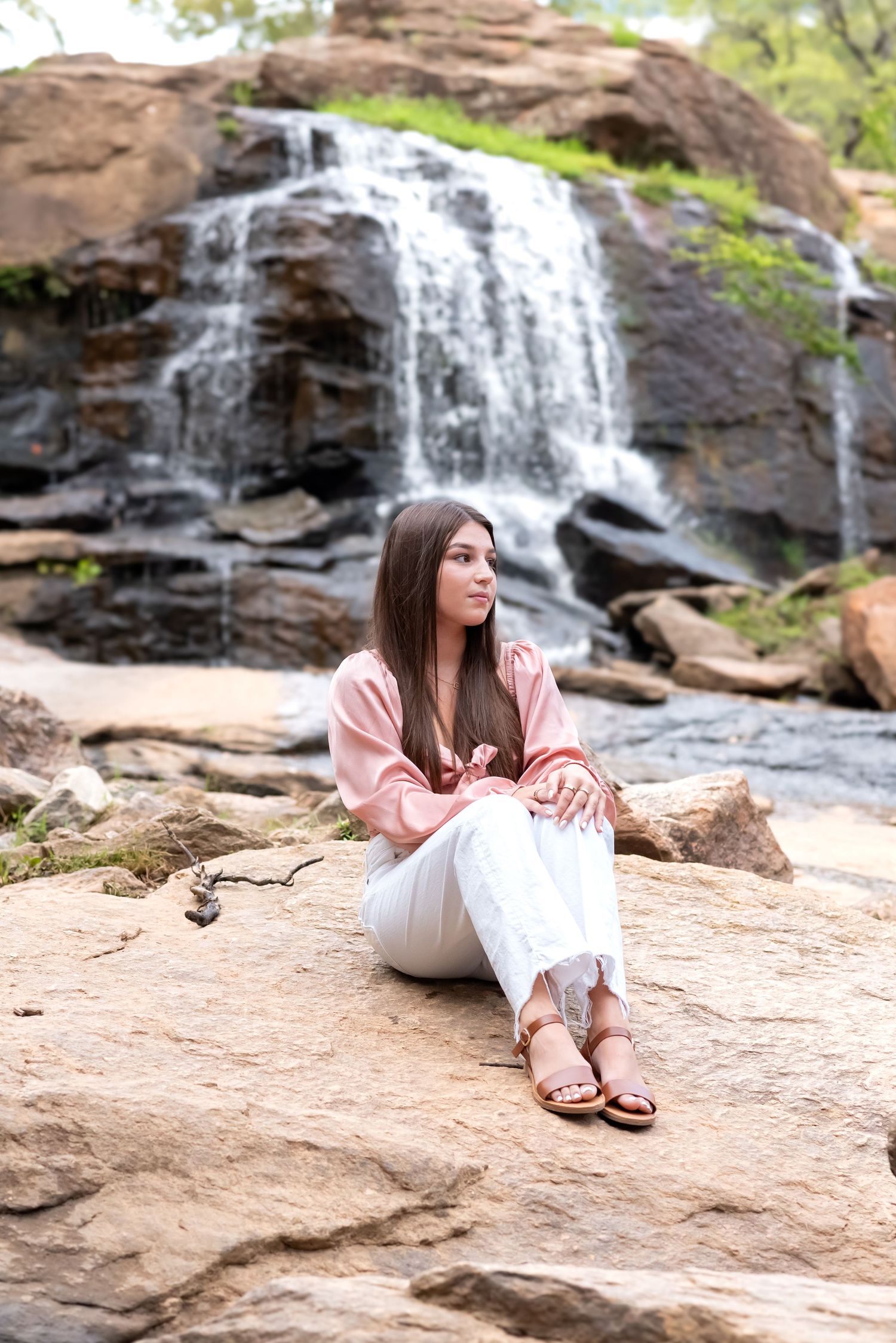 Young woman in pink blouse and white jeans sitting on rocks in front of a cascading waterfall.