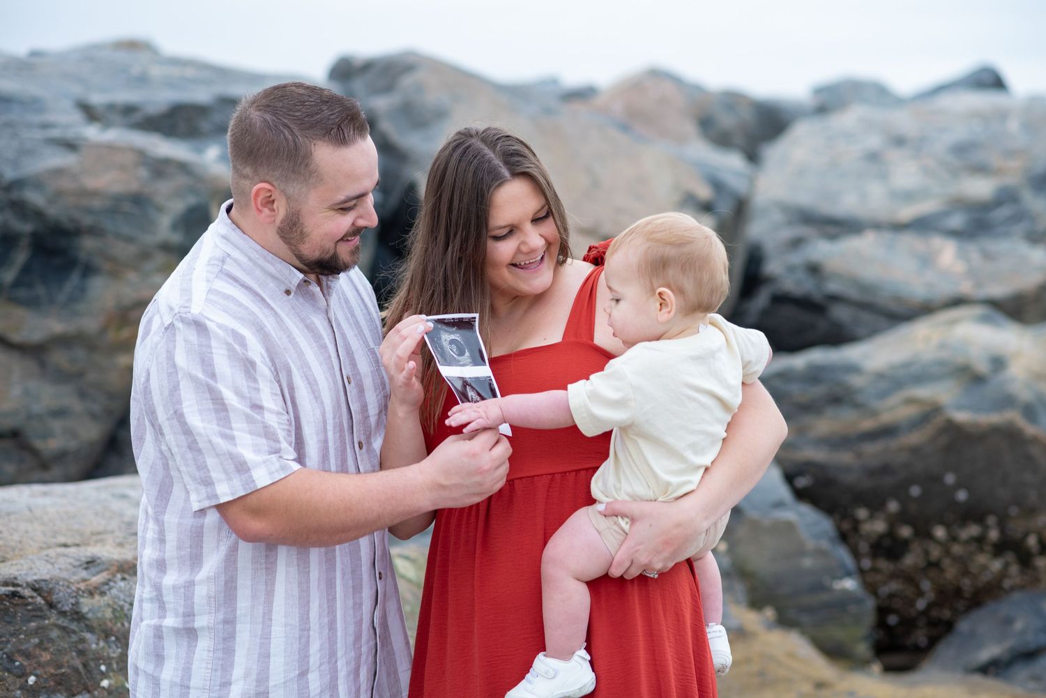 Family of three with baby looking at ultrasound photo on beach with rocky backdrop.