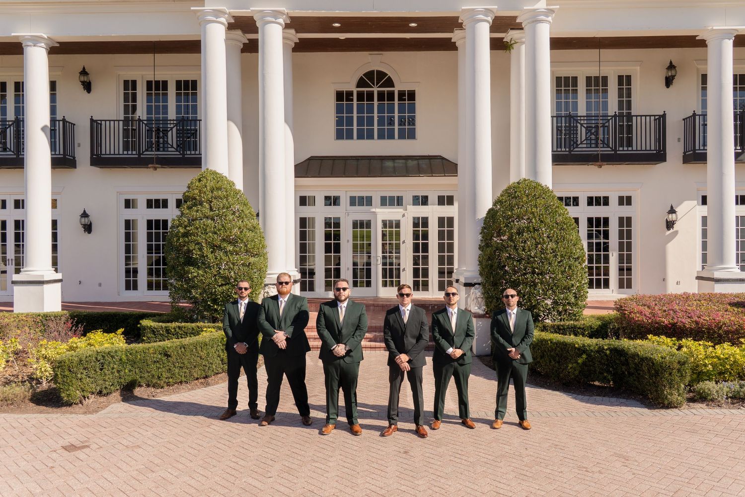 Six groomsmen in dark suits and sunglasses posing in front of an elegant white colonial wedding venue.
