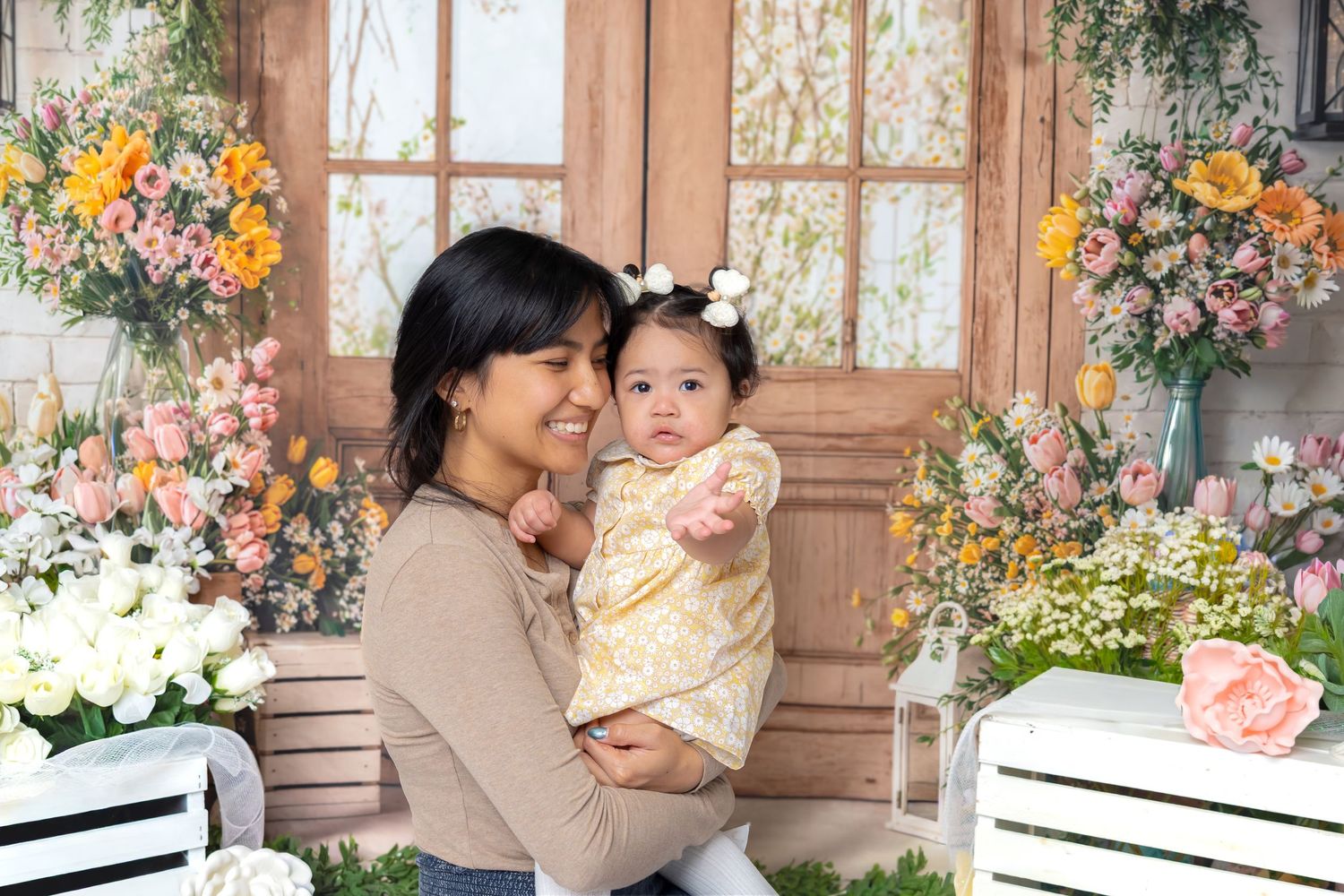 A woman holds a baby girl in a yellow floral outfit, posing in front of a colorful flower-decorated backdrop.