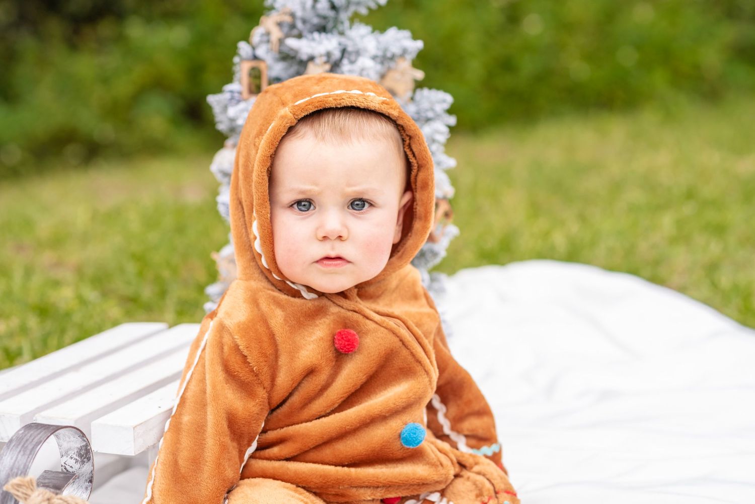Adorable baby in brown reindeer costume sitting on white blanket outdoors near Christmas tree.