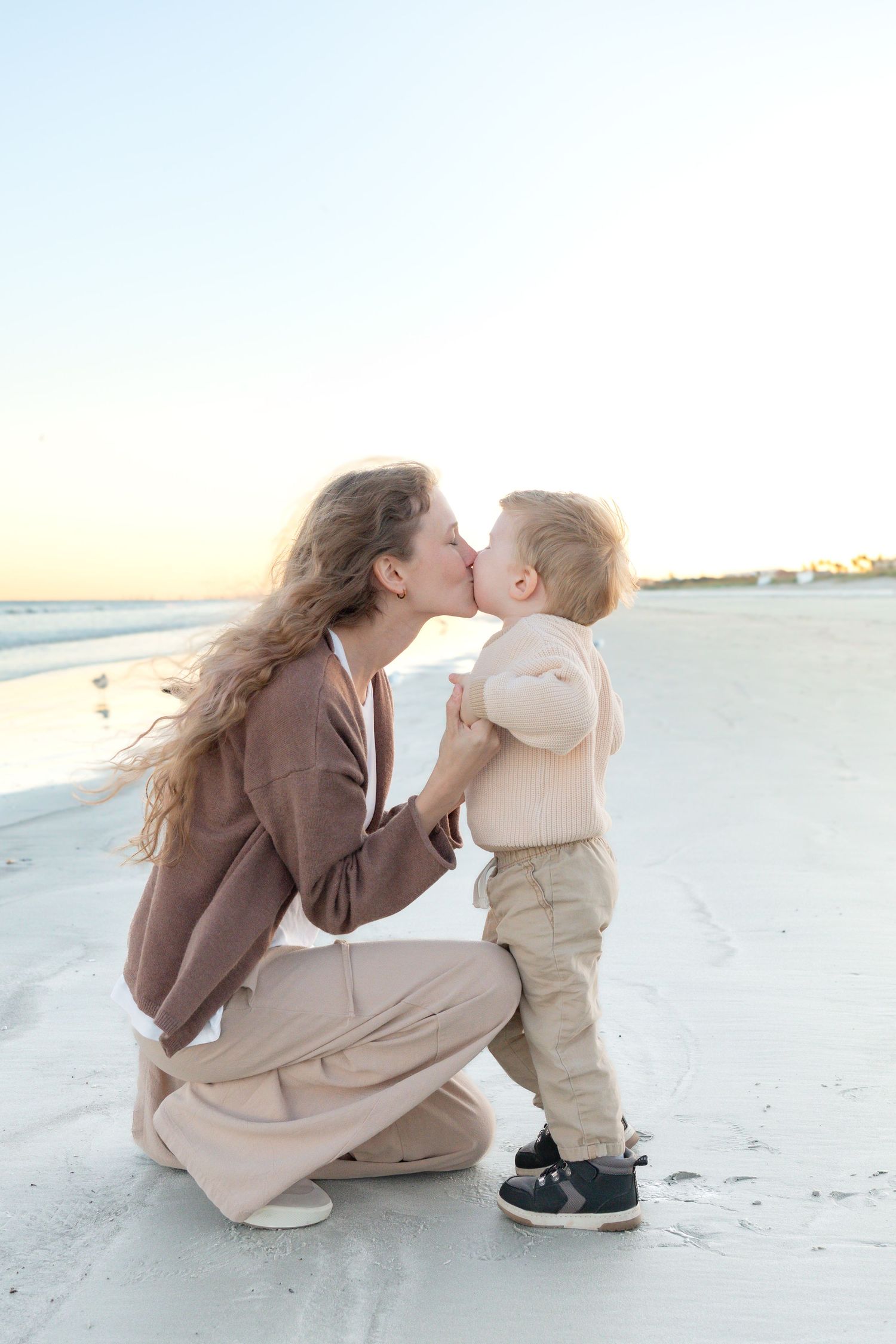 A mother kneels on a white sand beach, sharing a tender kiss with her toddler son at sunset.