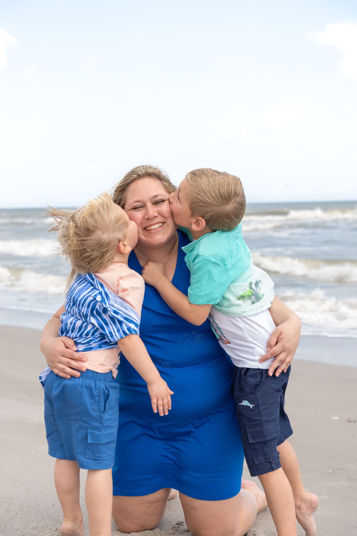 A smiling woman in blue kneels on a beach while two young boys give her kisses on her cheeks.
