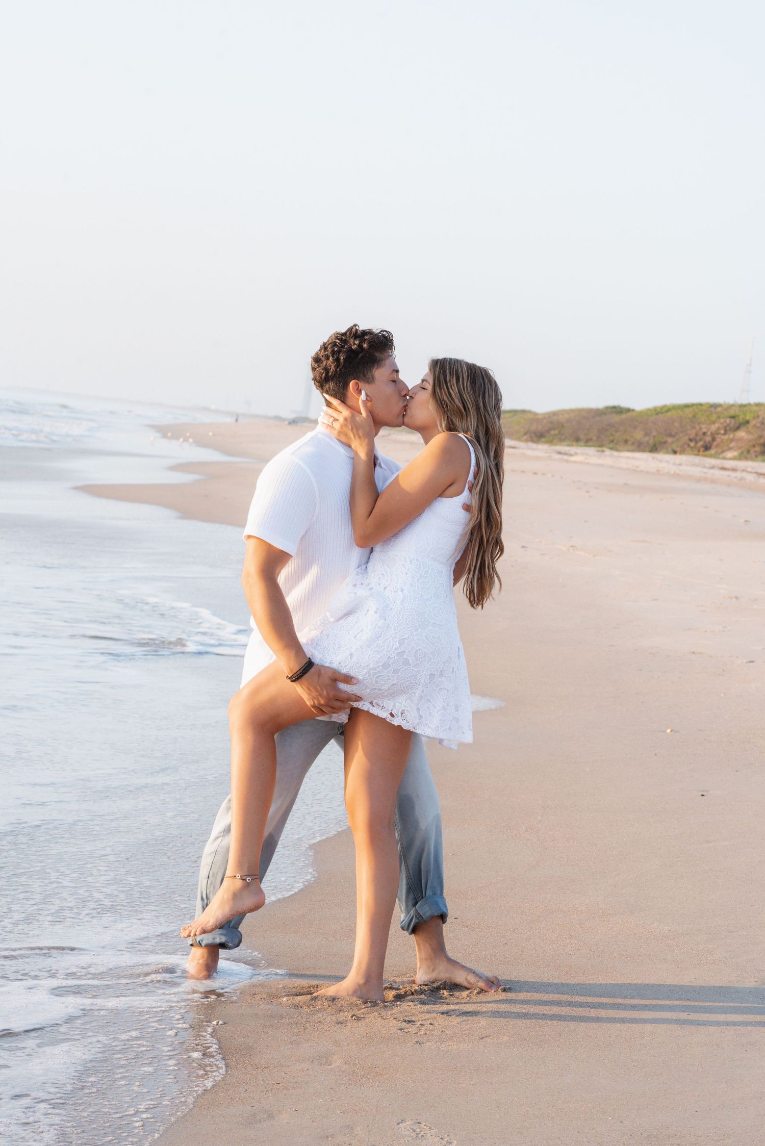 Couple sharing a romantic kiss on a sandy beach at sunset, woman in white dress lifted by man in white shirt and jeans.