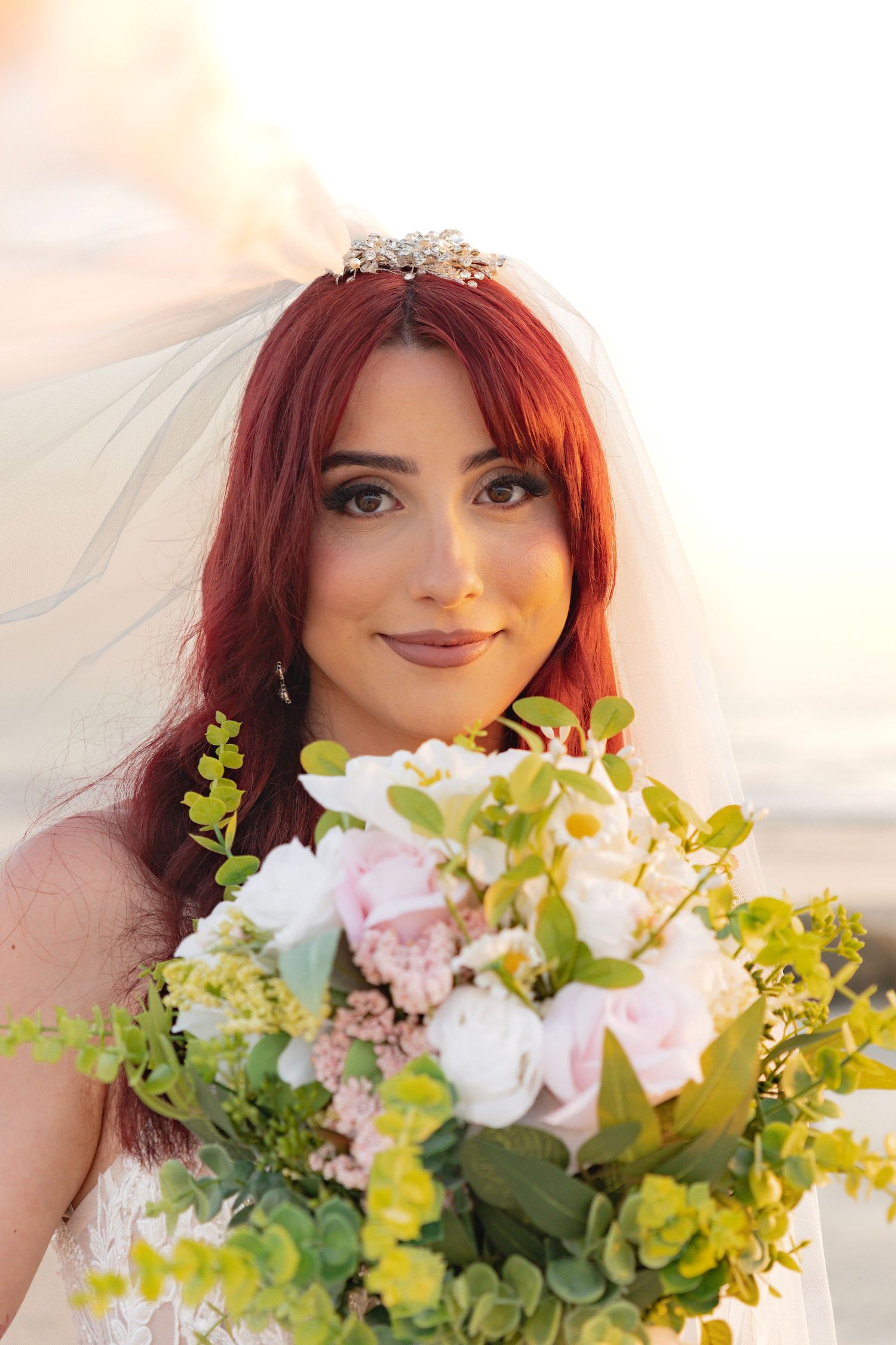 Red-haired bride wearing a tiara and veil holds a bouquet of white, pink, and green flowers outdoors.