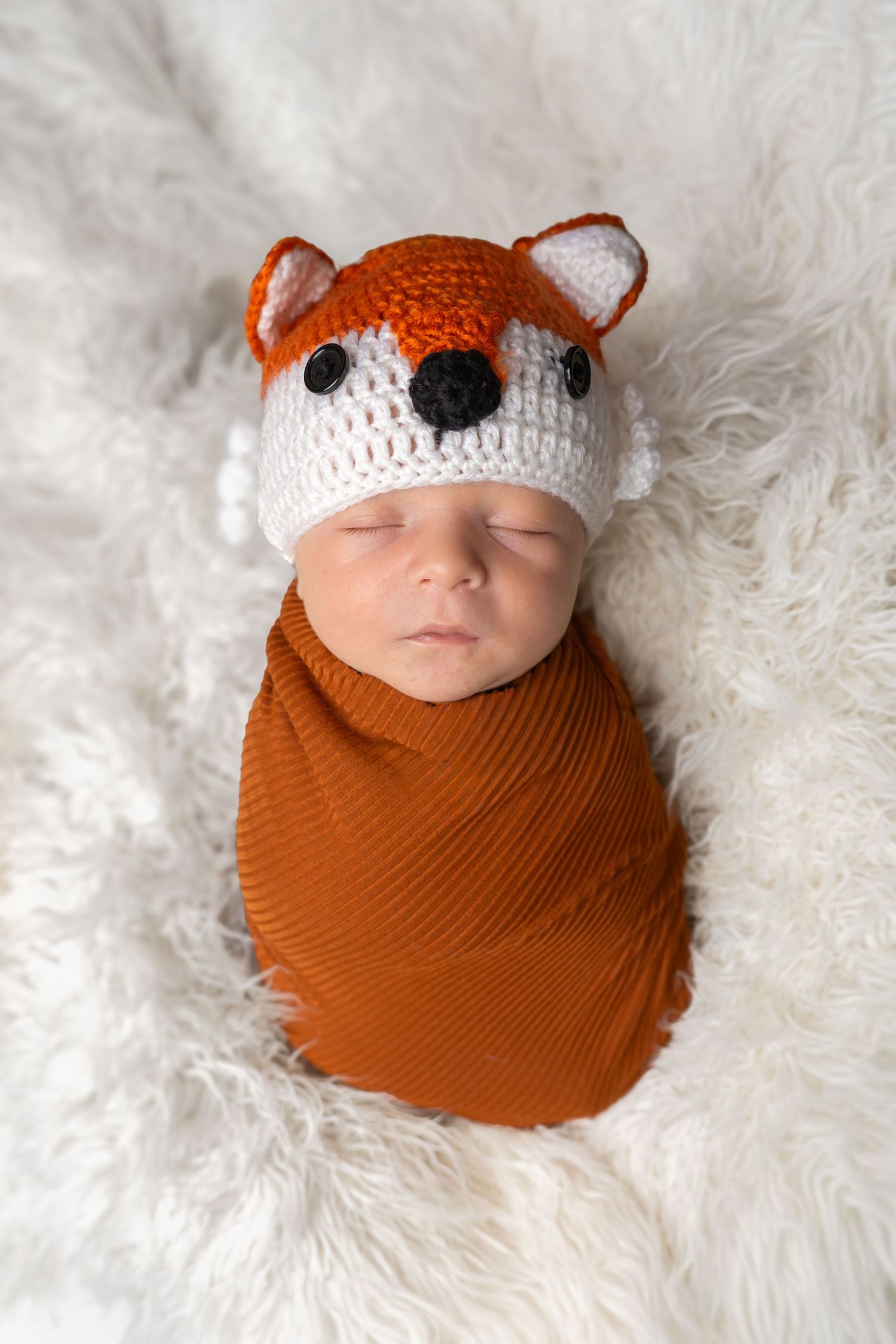 Sleeping newborn baby wrapped in orange swaddle wearing a crocheted fox hat on a white fluffy blanket.
