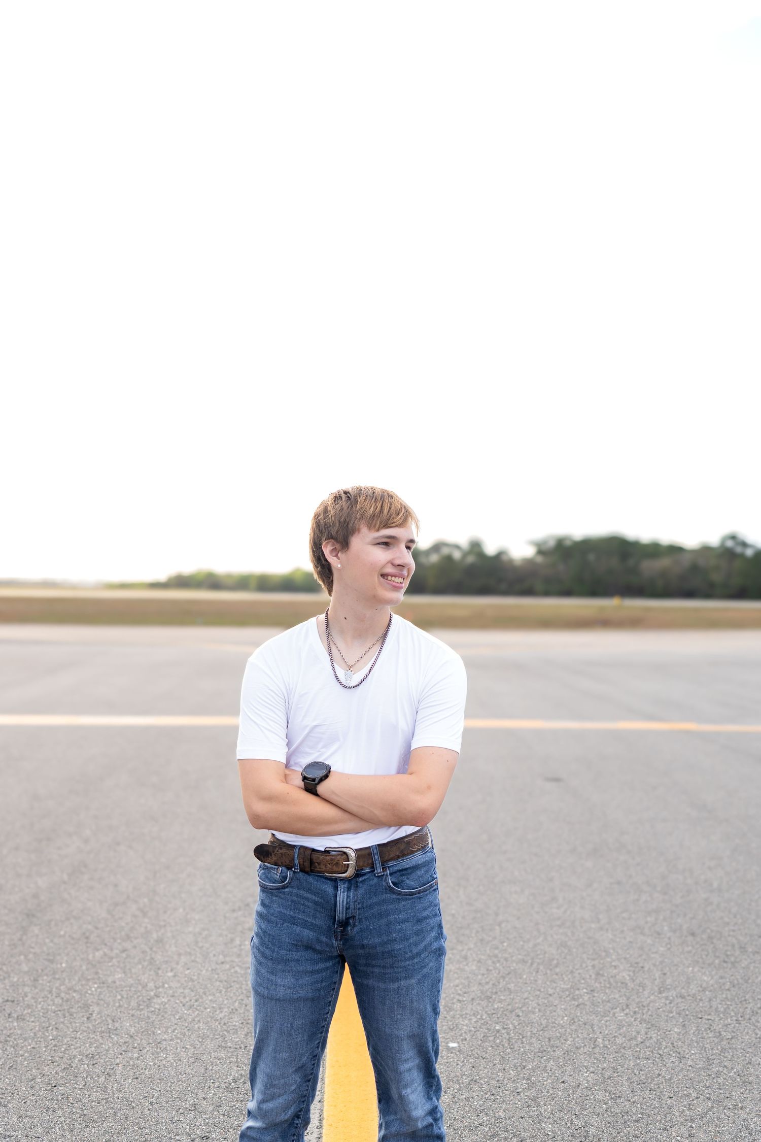 Smiling teenage boy in white t-shirt and jeans standing with arms crossed on an airport runway with yellow centerline.