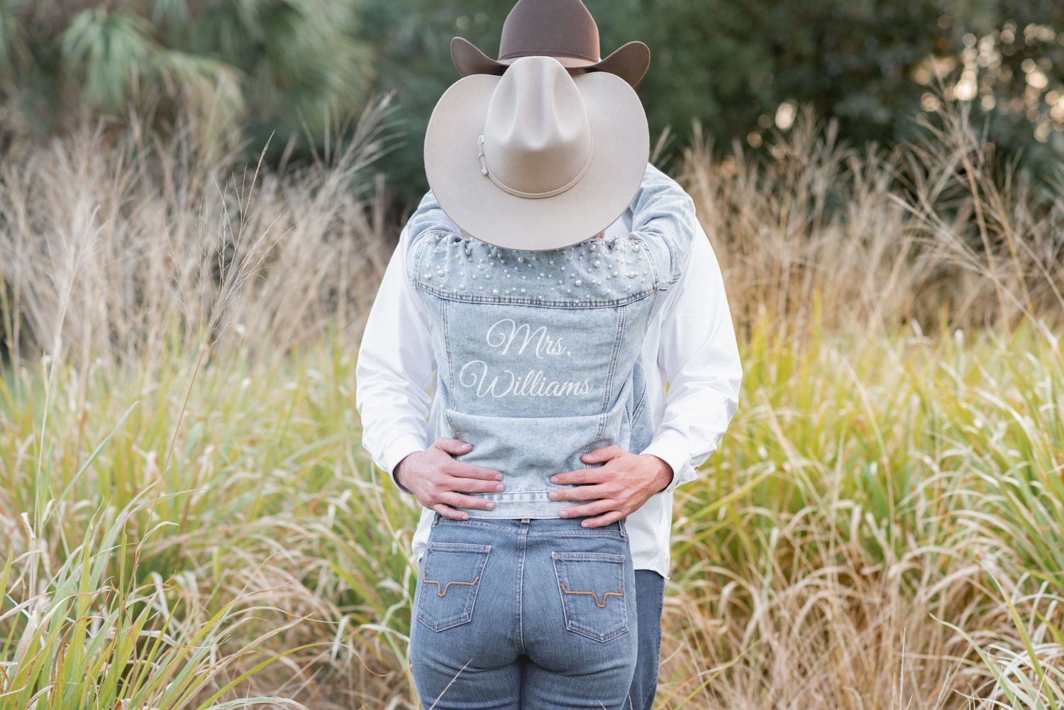 Person wearing a cowboy hat and denim vest with Mrs. Williams script, standing in a field of tall grass.