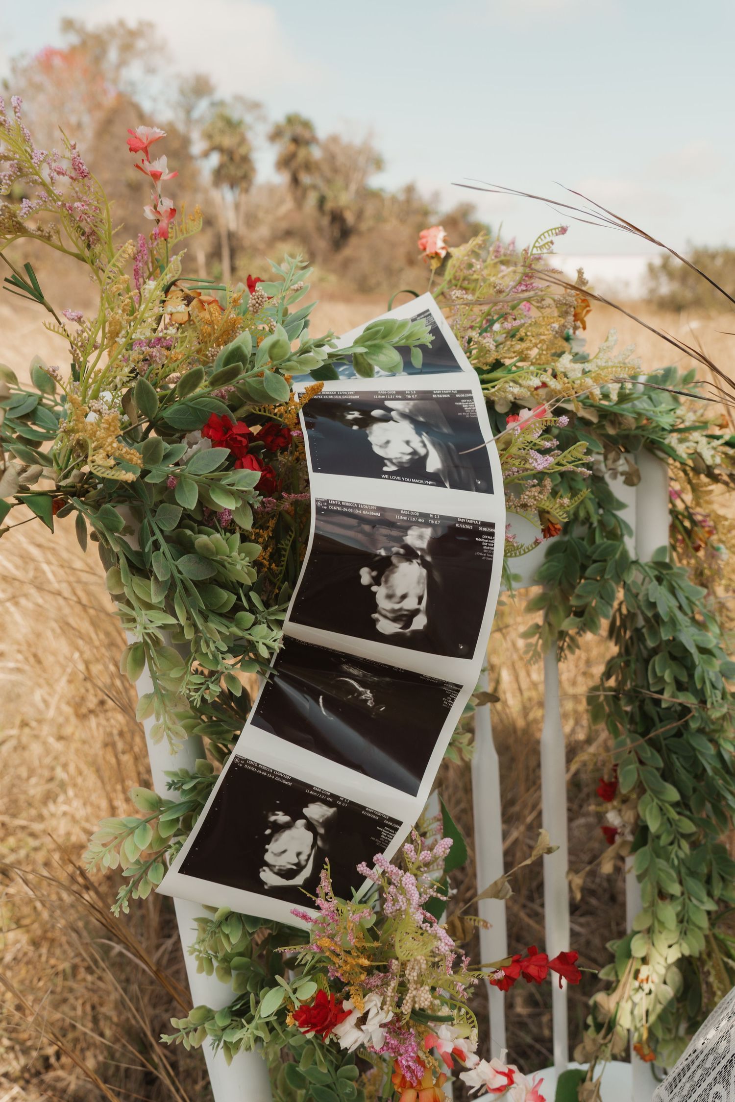 Ultrasound photos displayed on a floral arch with red and green flowers in an outdoor field setting.