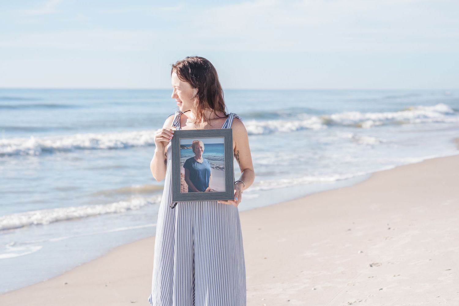 Woman in striped dress holding a framed photo of a man while standing on a sunny beach with ocean waves.