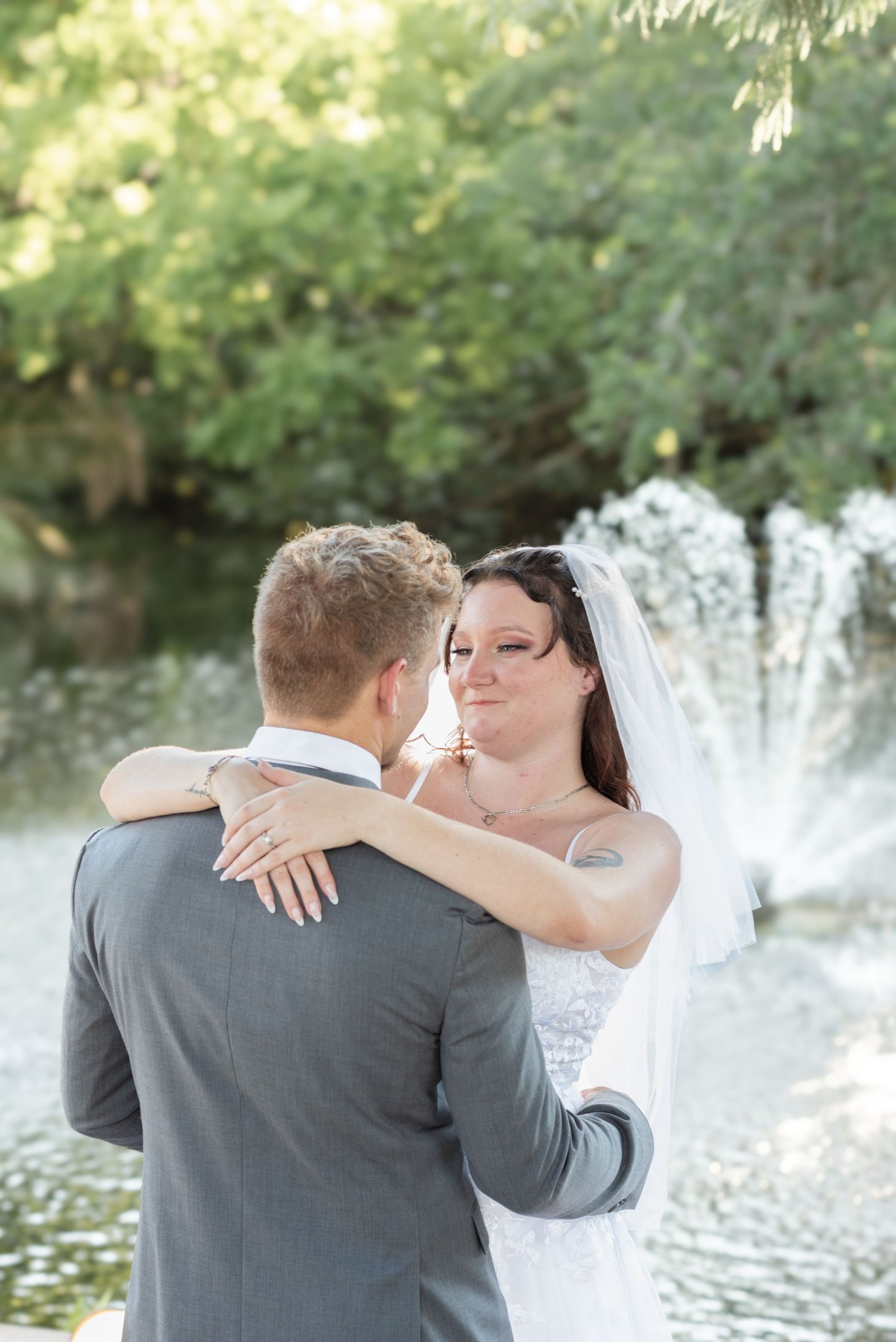 Bride in white gown and veil embraces groom in gray suit near a garden fountain on their wedding day.