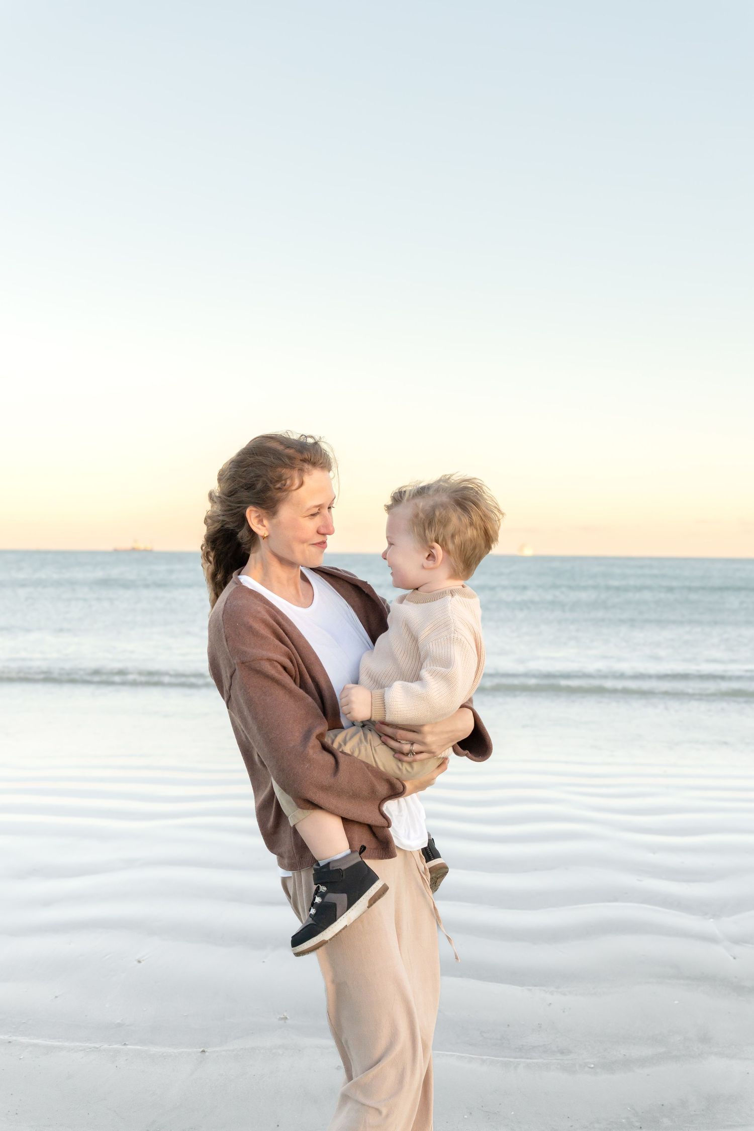 A woman holds a toddler on a serene beach at sunset, both sharing a tender moment by the ocean.