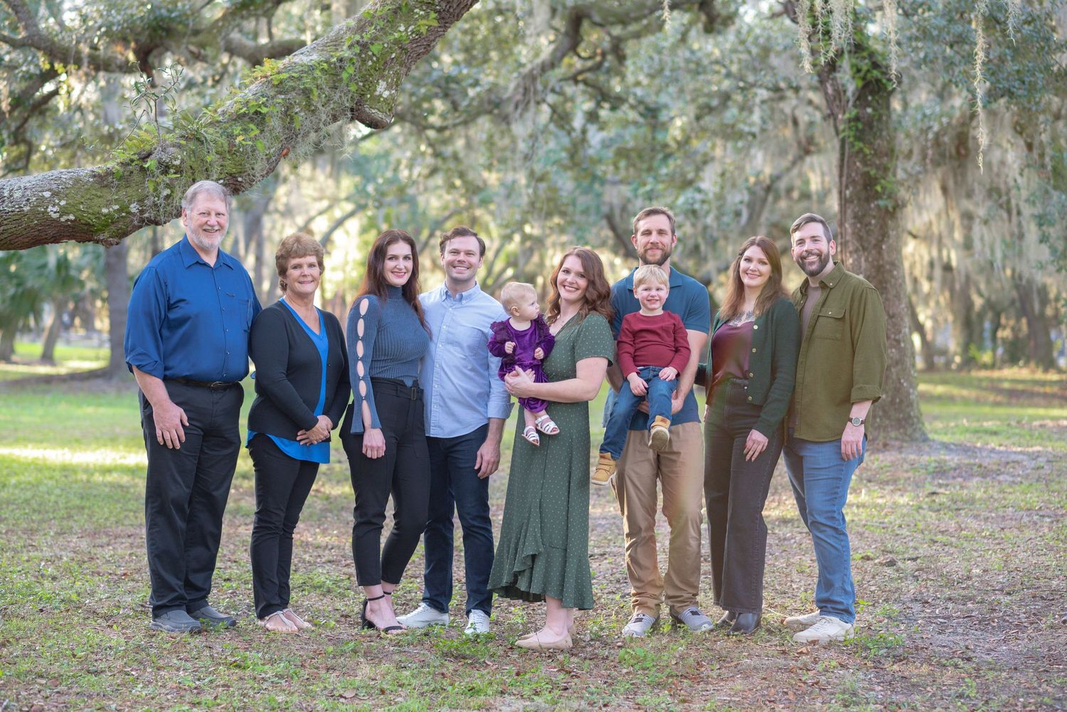 A joyful family group of eight poses outdoors under sprawling moss-covered oak trees in a lush green park setting.