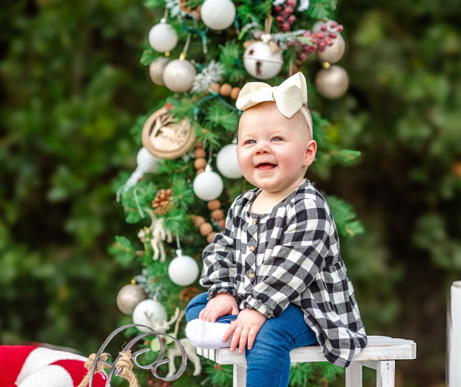 Happy baby girl in plaid shirt with gold bow sitting near decorated Christmas tree outdoors.
