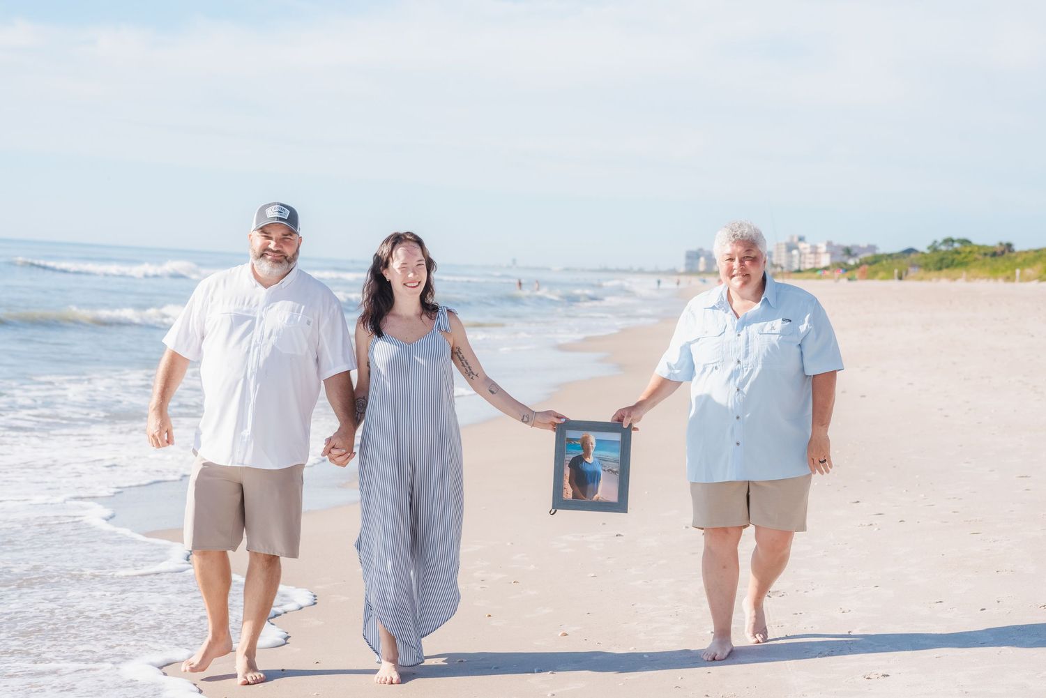 Three people walking on beach holding framed photo as memorial tribute.