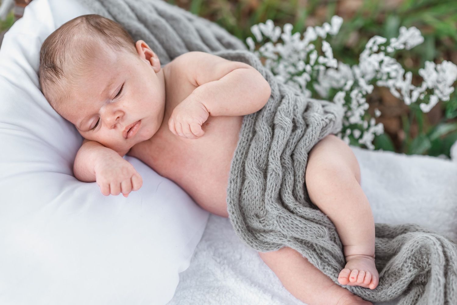 Peaceful newborn baby sleeping on white cushion wrapped in soft gray knit blanket.